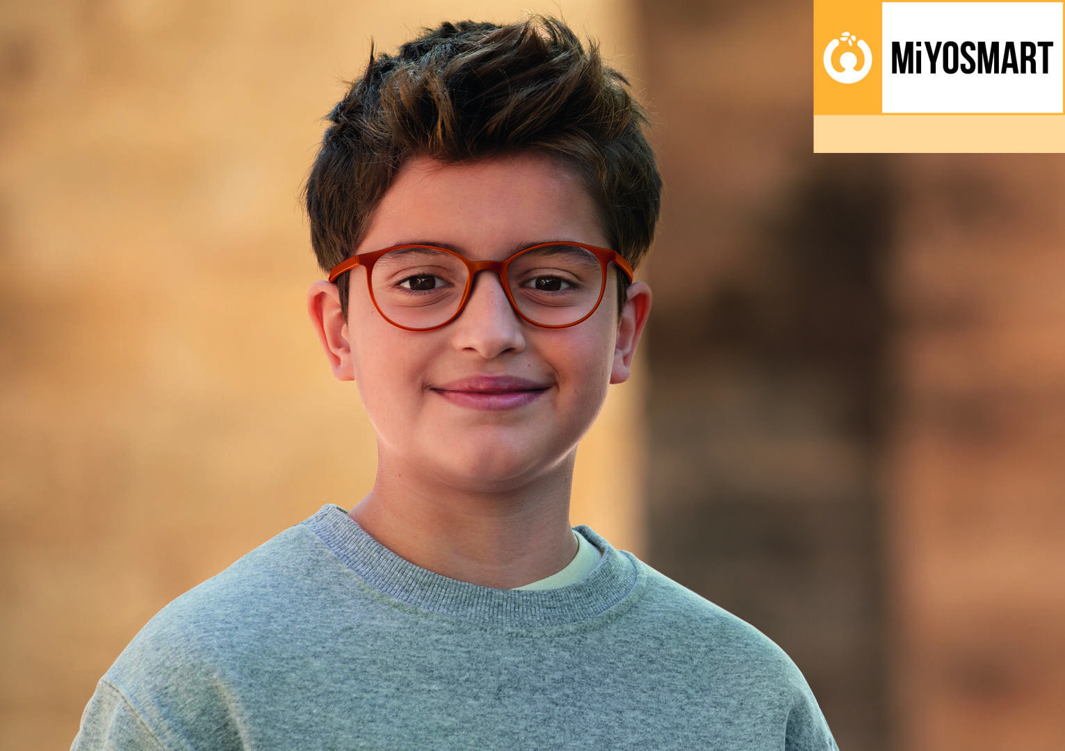 Ein lächelnder Junge mit kurzen braunen Haaren und Brille, der ein graues Sweatshirt trägt, steht im Freien vor einem unscharfen Hintergrund. Das MiYOSMART-Logo erscheint in der oberen rechten Ecke.