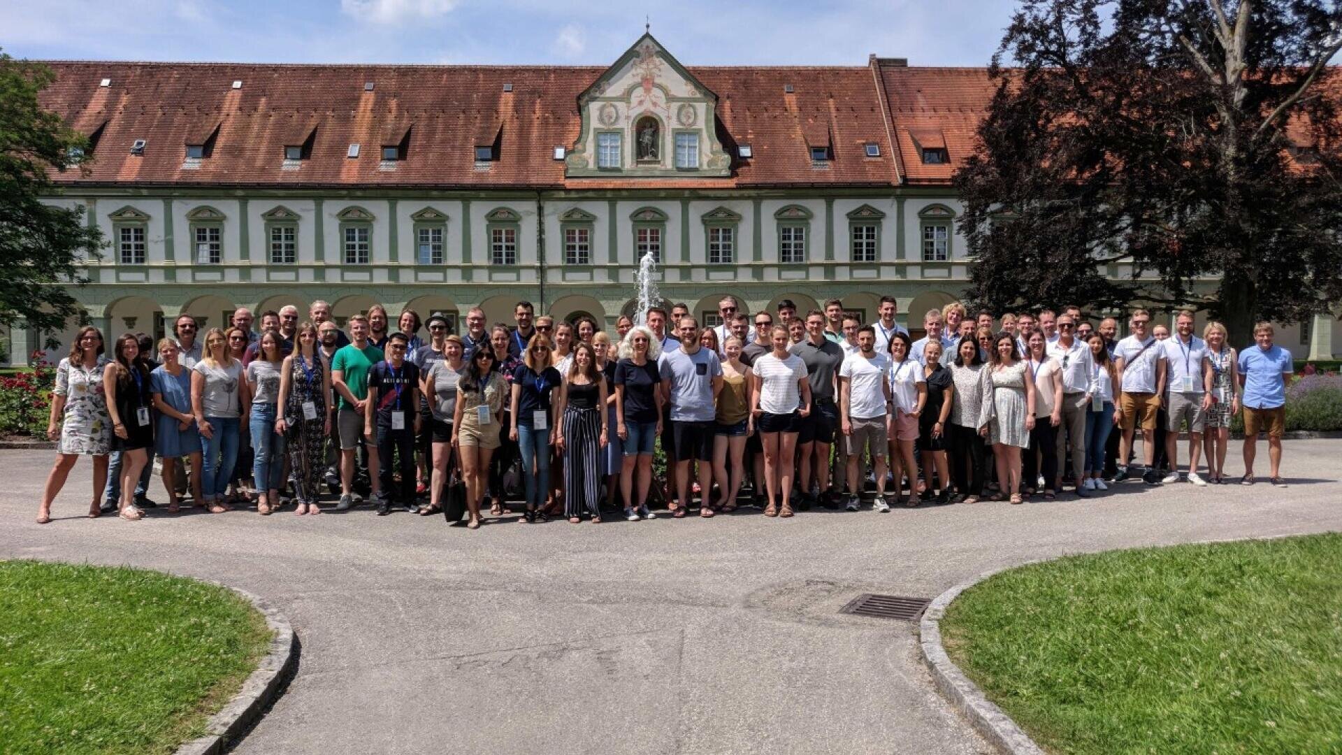 A large group of people stand together for a group photo in front of a historic building with a red-tiled roof and arched windows on a sunny day. Trees and grass frame the paved courtyard.