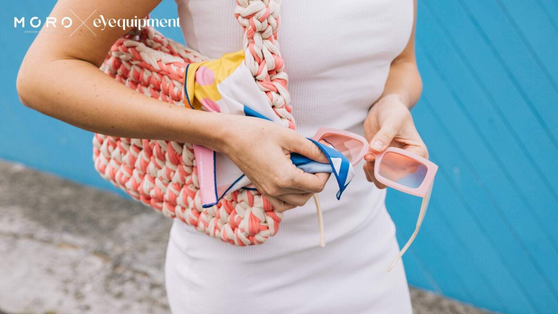 A person in a white dress holds pink sunglasses and a colorful scarf, with a woven pink and white bag over their shoulder, standing in front of a blue wall. The Moro Eyequipment logo is in the top left corner.