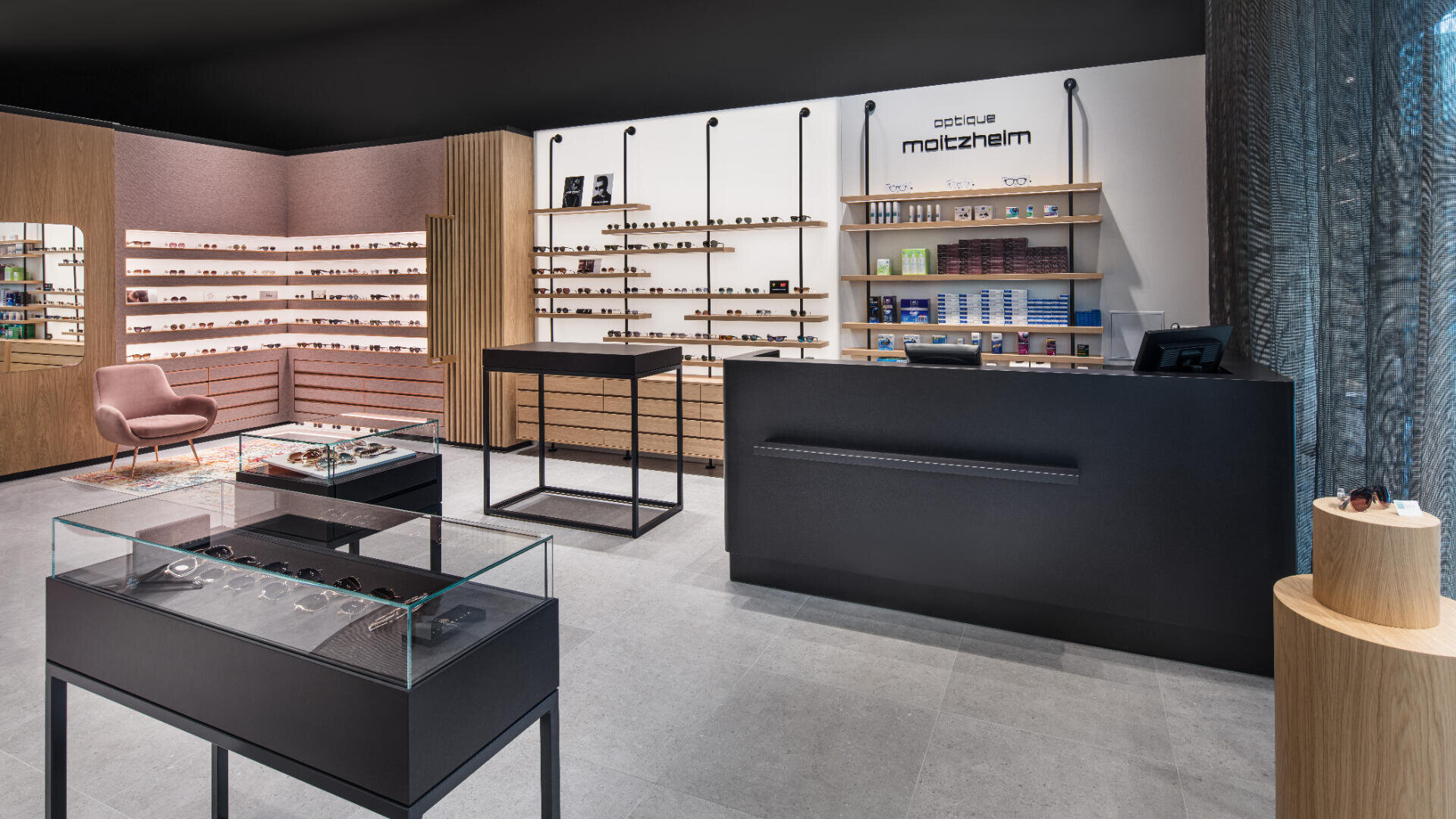 Modernes Interieur eines Brillengeschäfts mit Holzregalen, auf denen verschiedene Brillen ausgestellt sind, schwarzen Vitrinen, einer schwarzen Theke, einem einzelnen rosa Stuhl und minimalistischem Dekor in einem sauberen, organisierten Raum.