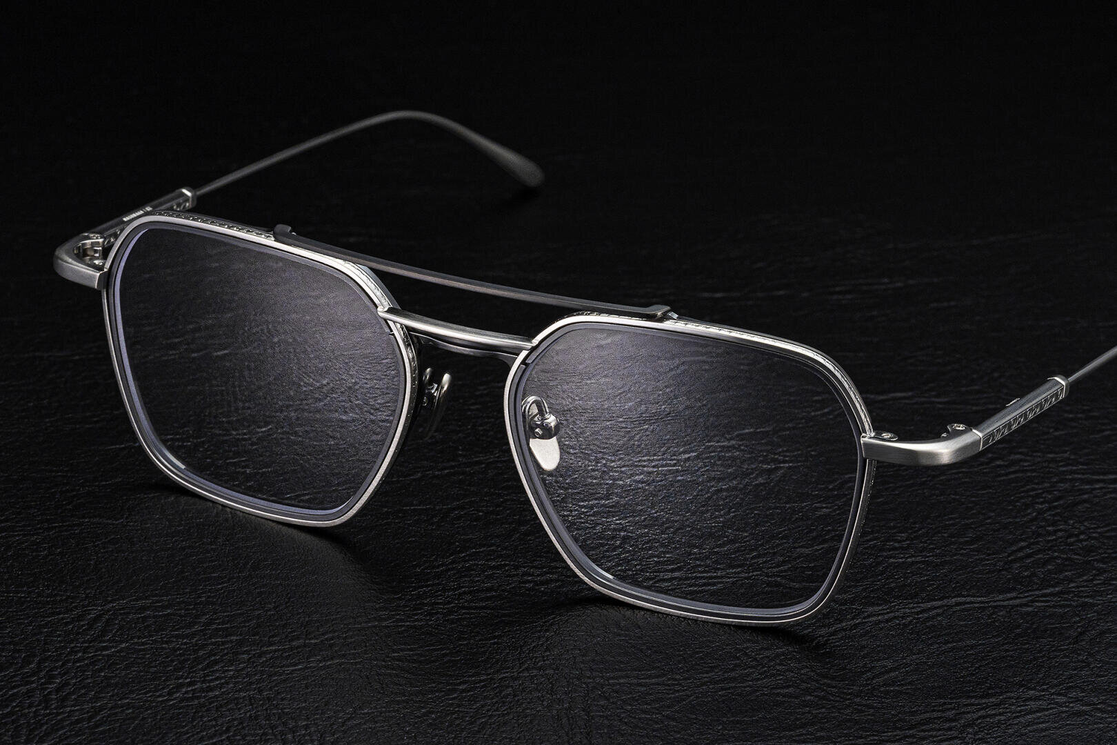 A pair of stylish, silver metal eyeglasses with large, rectangular lenses and thin arms, displayed on a black textured surface.