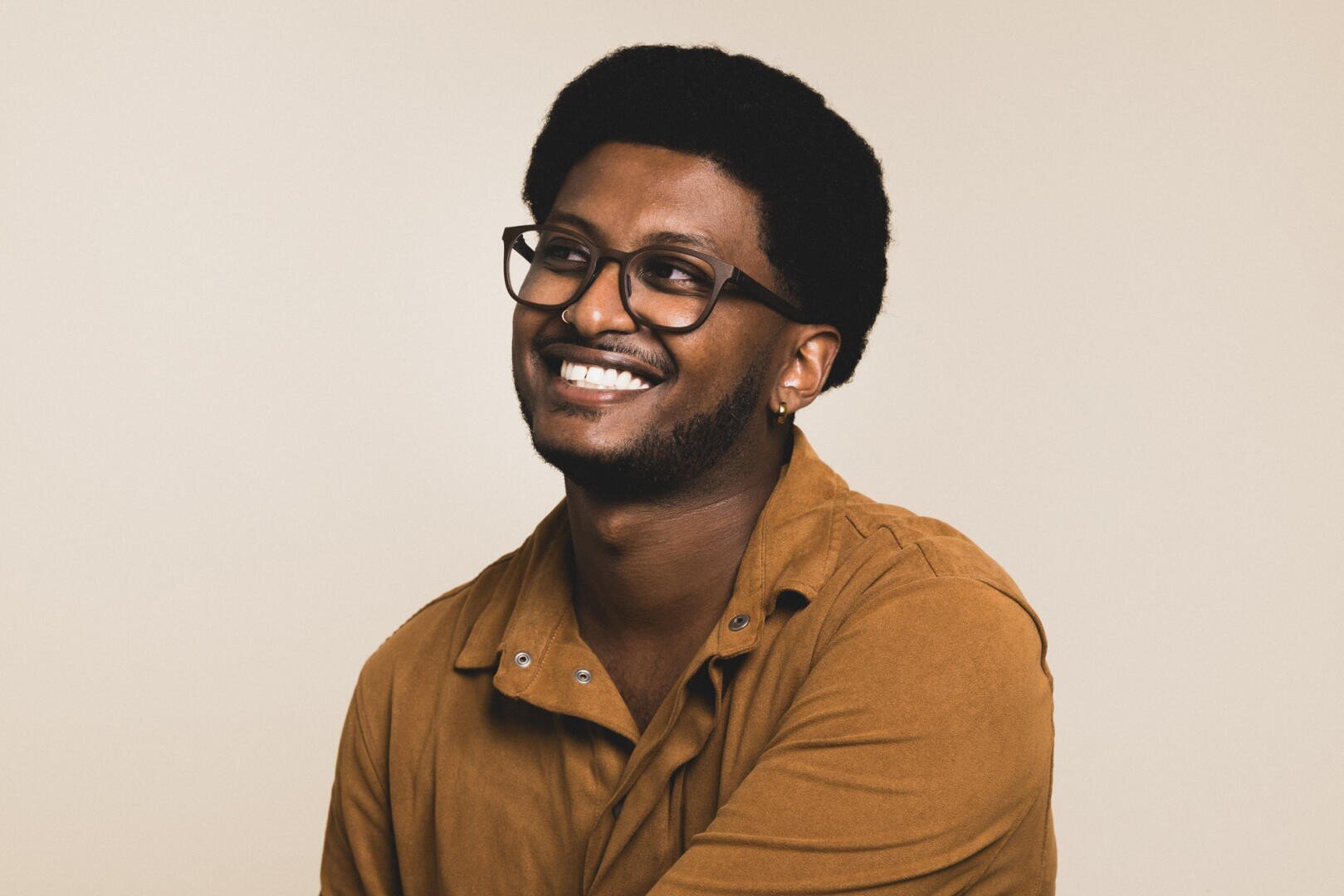 Ein lächelnder Mann mit dunklem lockigem Haar, schwarzer Brille und braunem Hemd sitzt vor einem schlichten hellen Hintergrund.