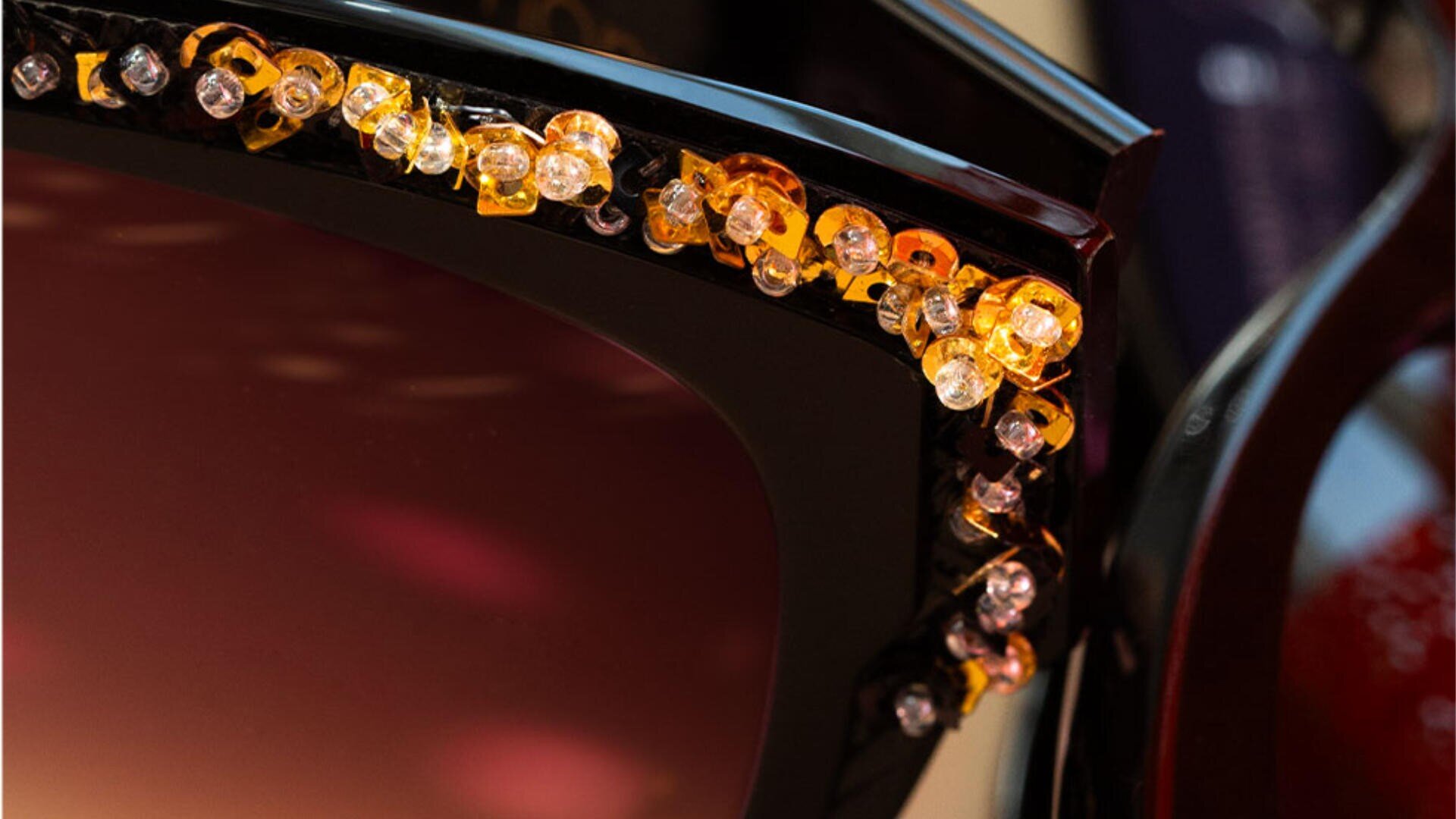 Nahaufnahme der Ecke einer dunklen Sonnenbrille, deren Rahmen mit kleinen bernsteinfarbenen, gelben und klaren Edelsteinen verziert ist.