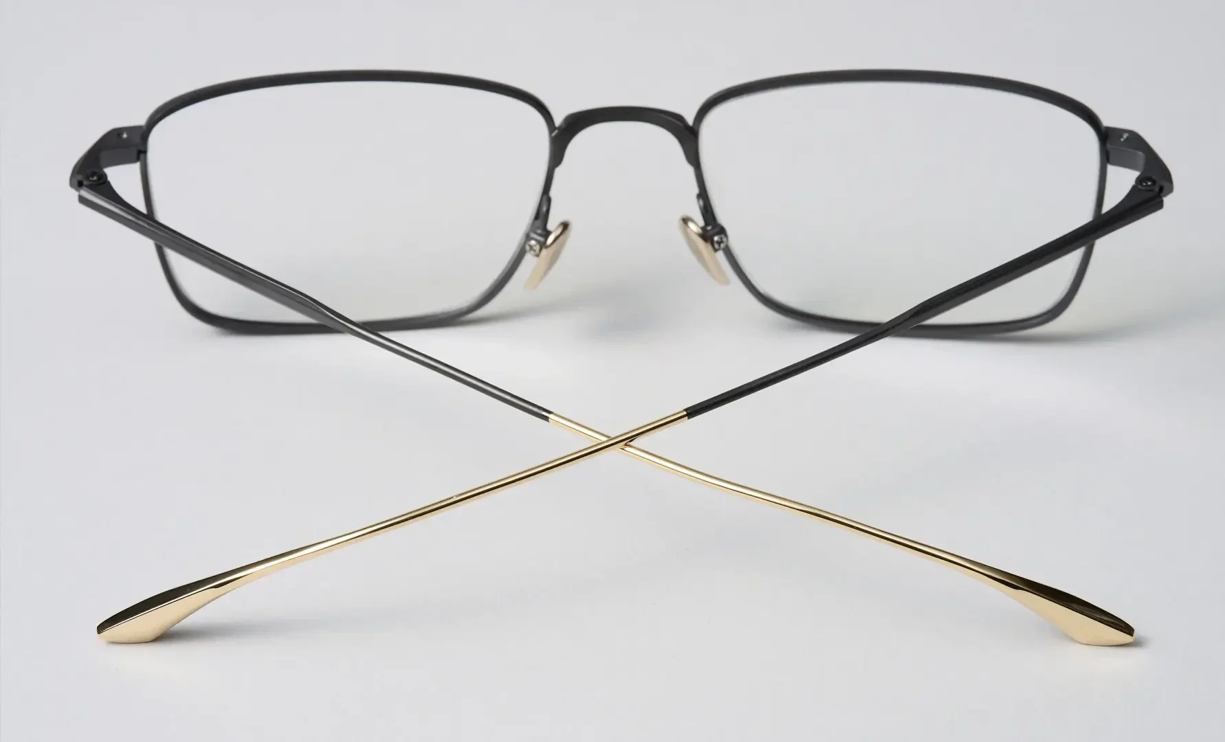 A pair of eyeglasses with black rectangular frames and gold temples, shown on a white surface with the temples crossed in an X shape in front of the frames.