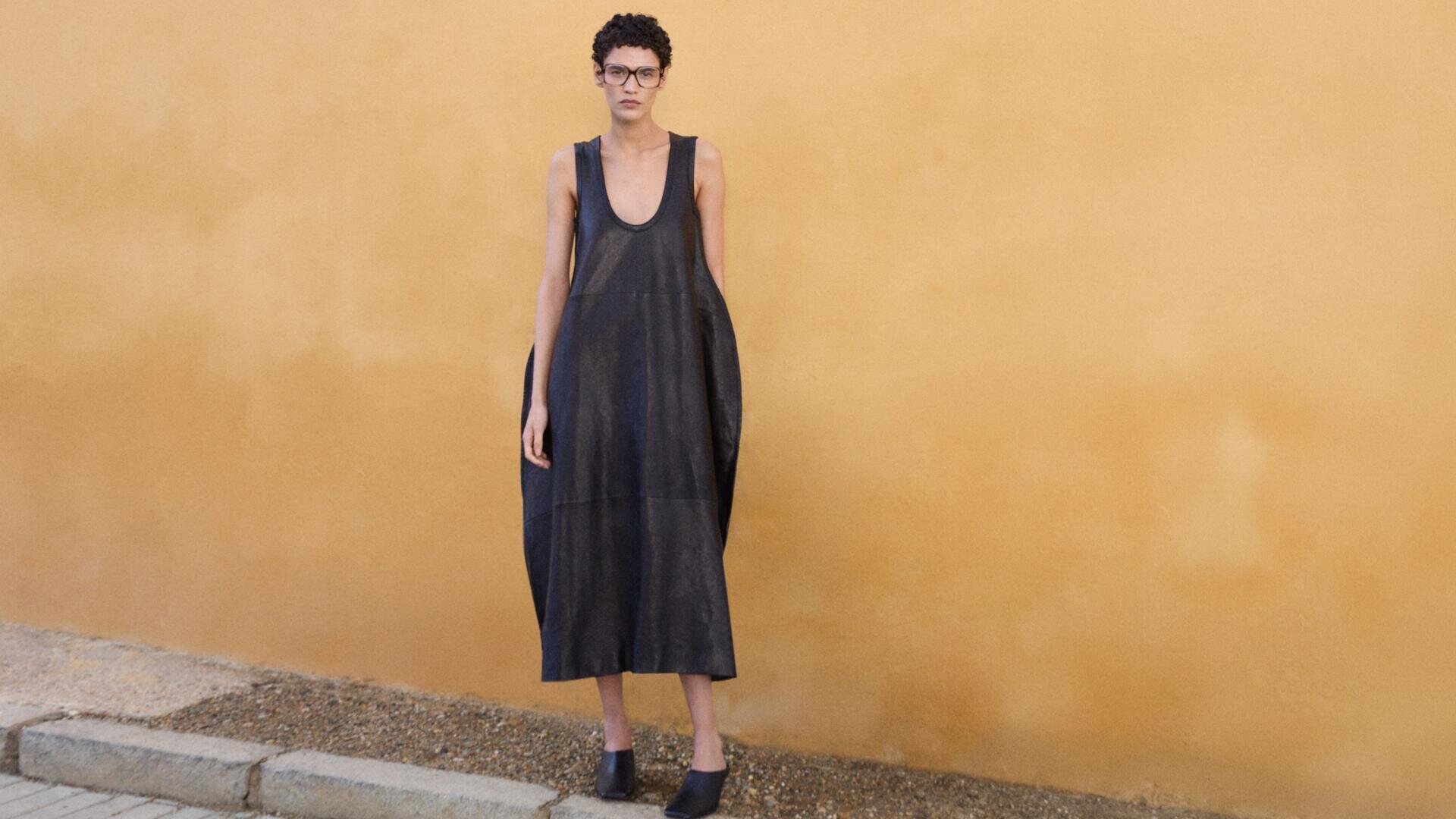A person with short curly hair and glasses stands against a mustard yellow wall, wearing a loose, sleeveless, dark gray dress and black shoes on a paved sidewalk.