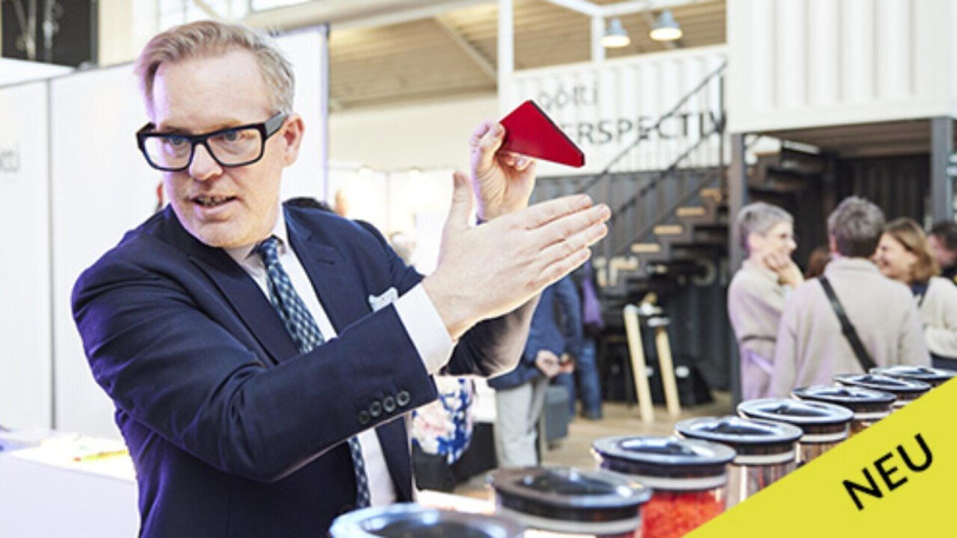 A man in a suit gestures while holding a red object, standing beside jars at an indoor event. Several people are gathered in the background. A yellow label with NEU is in the corner.