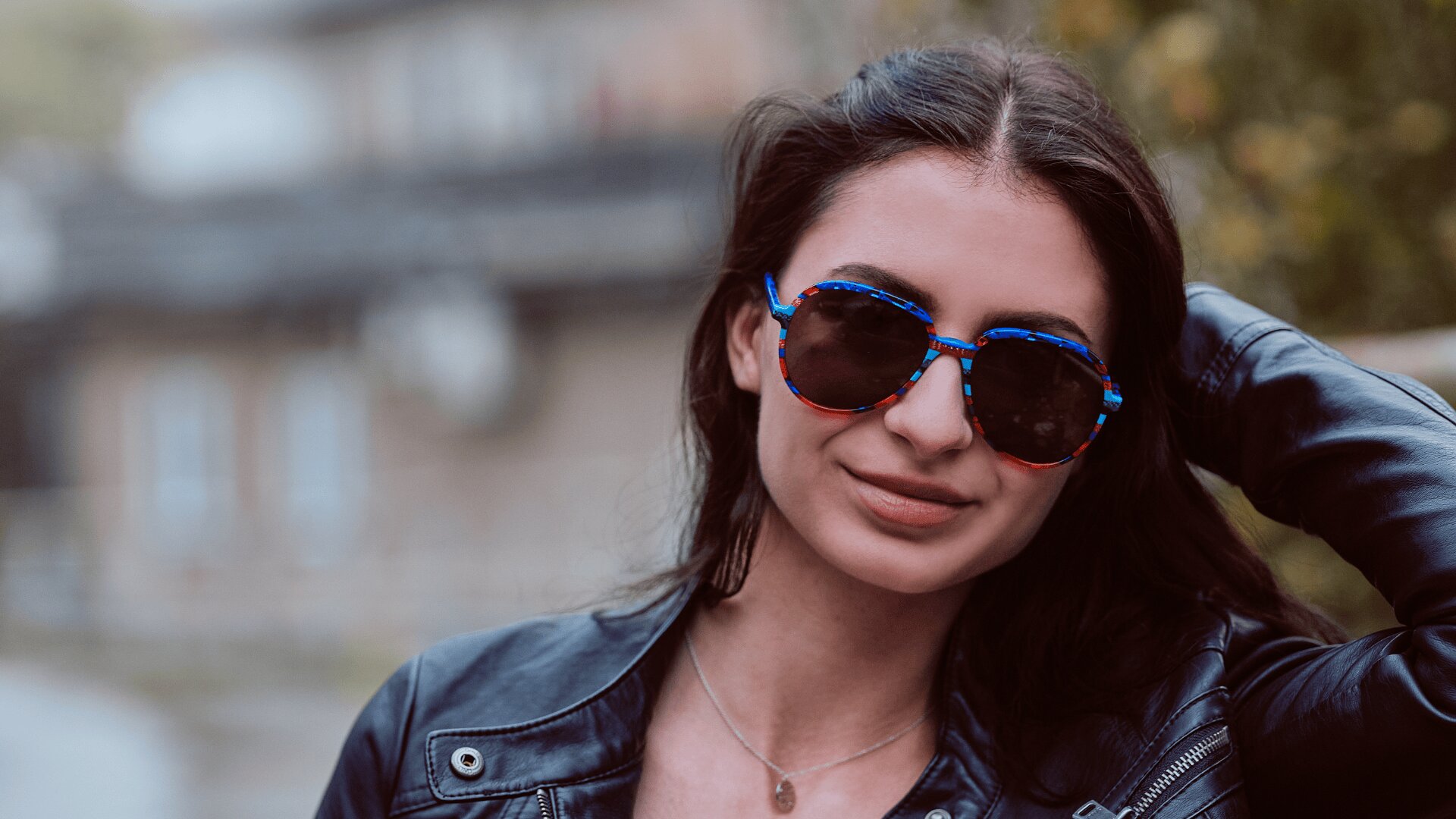 Eine Frau mit langen dunklen Haaren, die eine runde blaue und rote Sonnenbrille, eine schwarze Lederjacke und eine Halskette trägt, lächelt, während sie im Freien vor einem unscharfen Hintergrund ihre Hand auf ihren Kopf stützt.