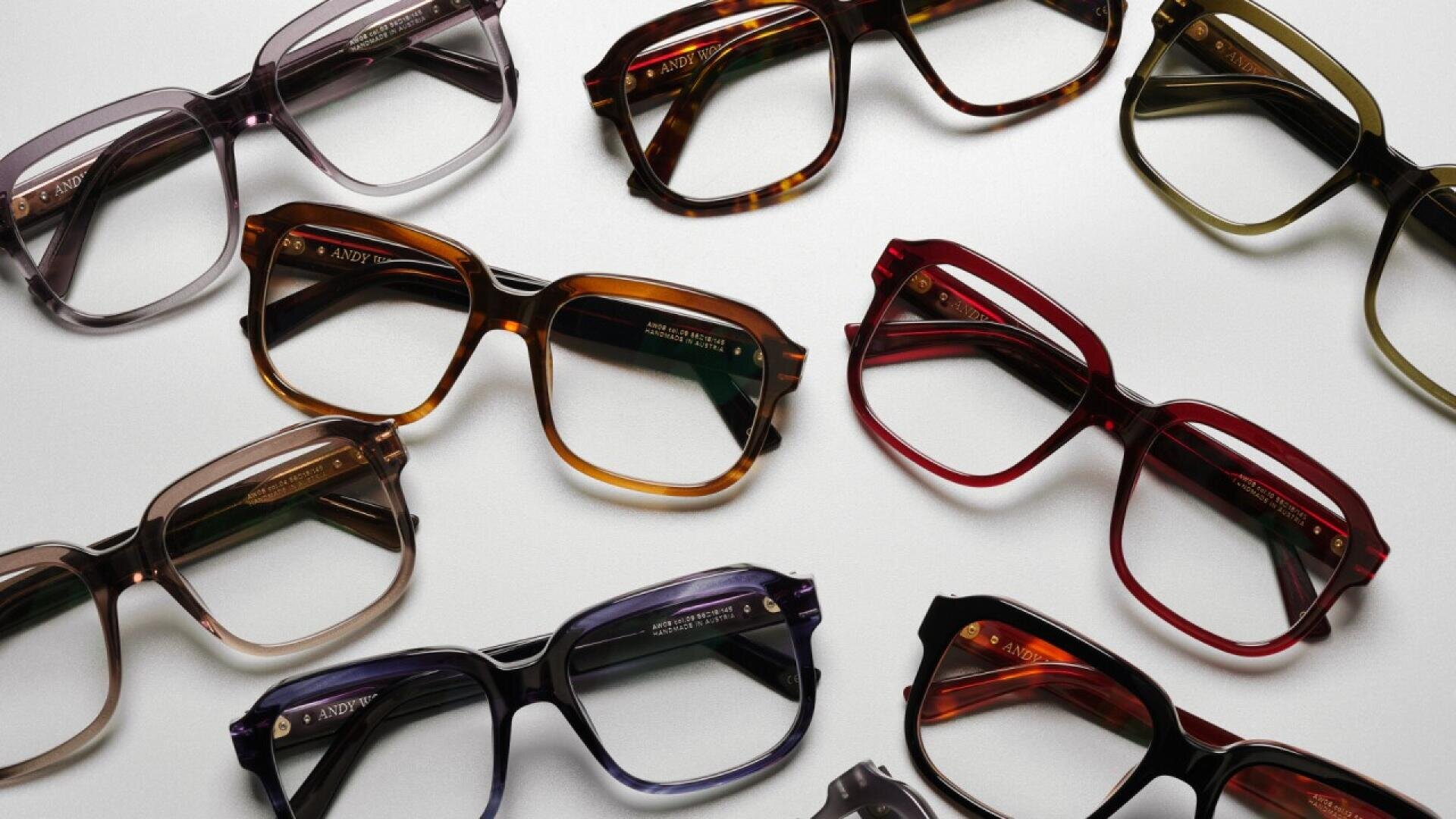 Several pairs of eyeglasses in various colors and styles are arranged on a flat white surface, showcasing their frames in shades of brown, black, gray, red, and purple.
