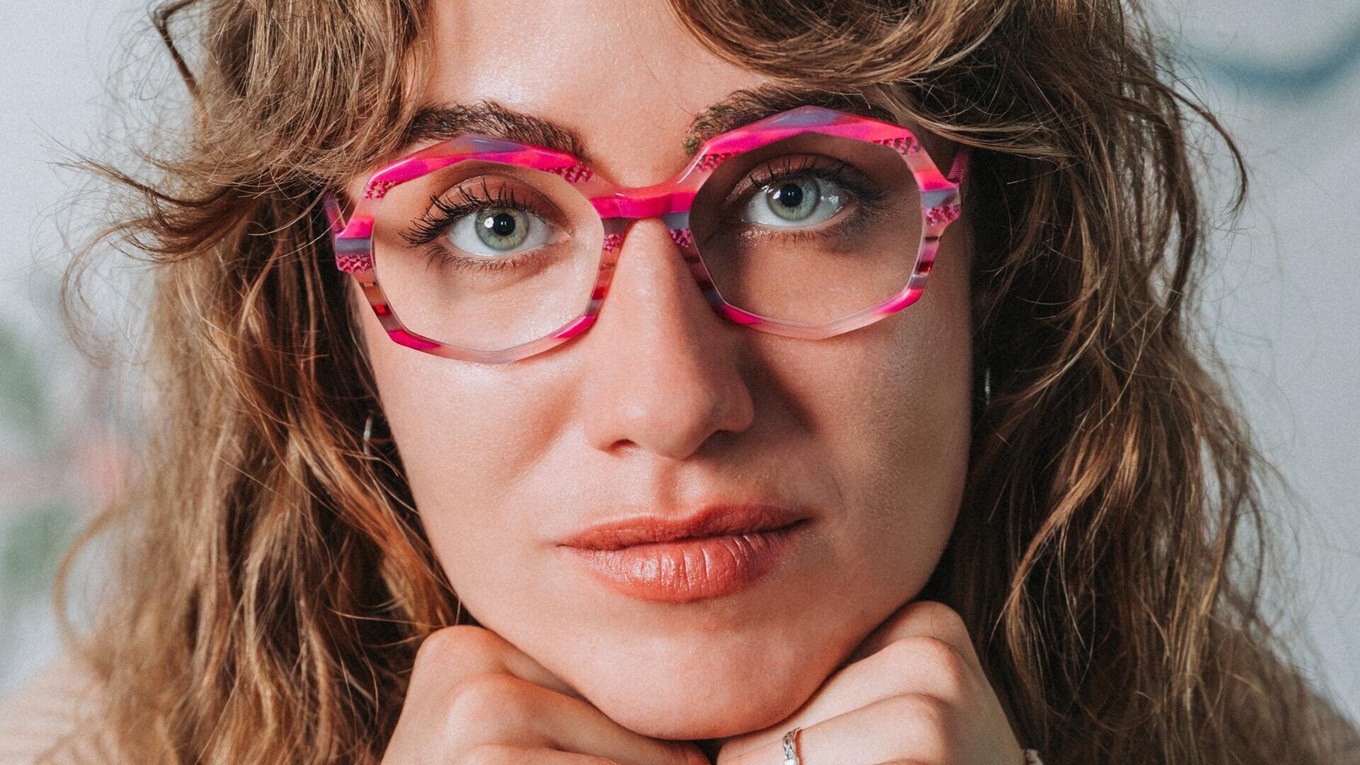 A person with wavy light brown hair and blue eyes looks directly into the camera, wears colorful, pink and purple patterned glasses and rests her chin on her hands.