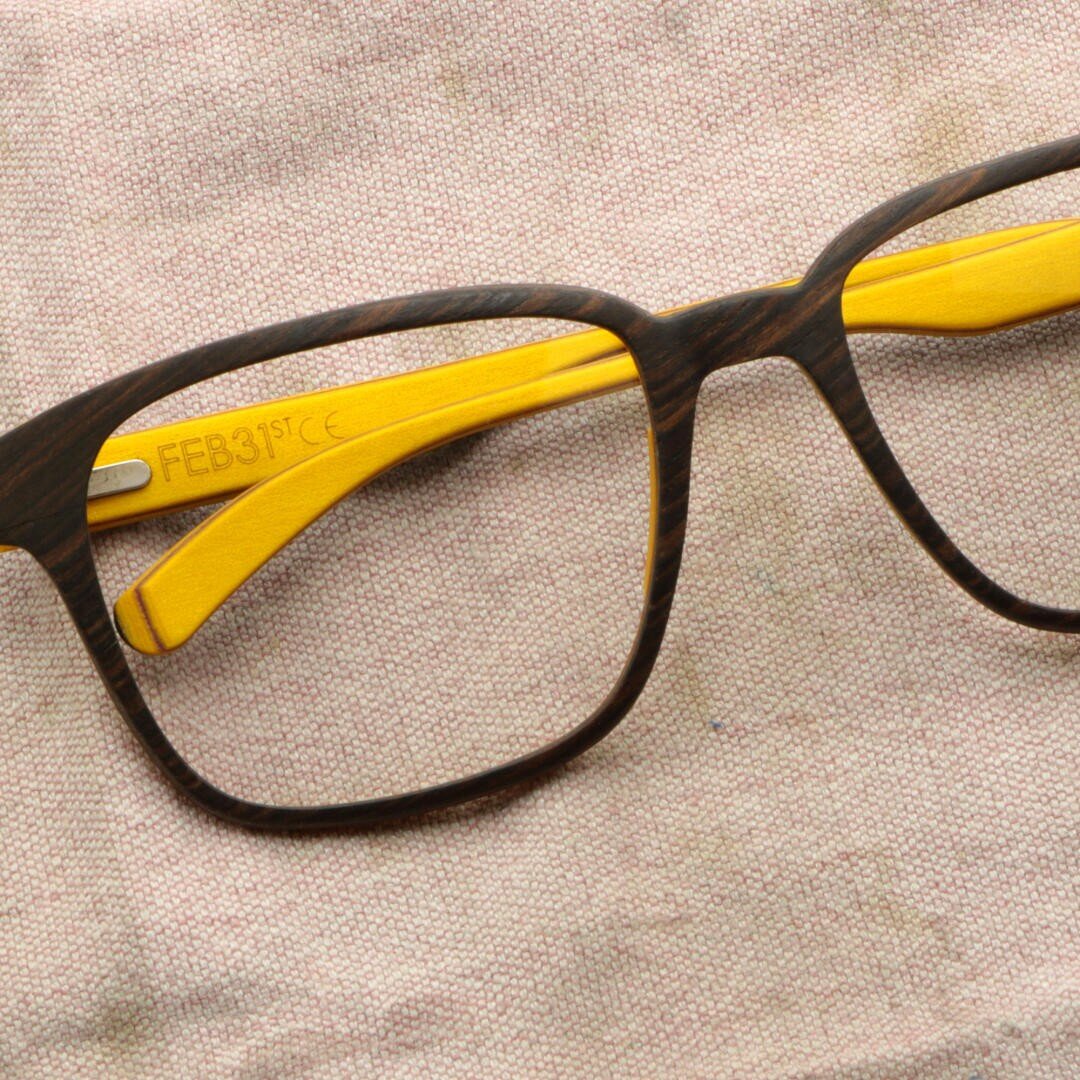 Eine Brille mit dunklem Holzrahmen und leuchtend gelben Bügeln, die auf einer hellen, strukturierten Stoffoberfläche ruhen. Die Innenseite eines Bügels ist mit FEB31st CE gekennzeichnet.