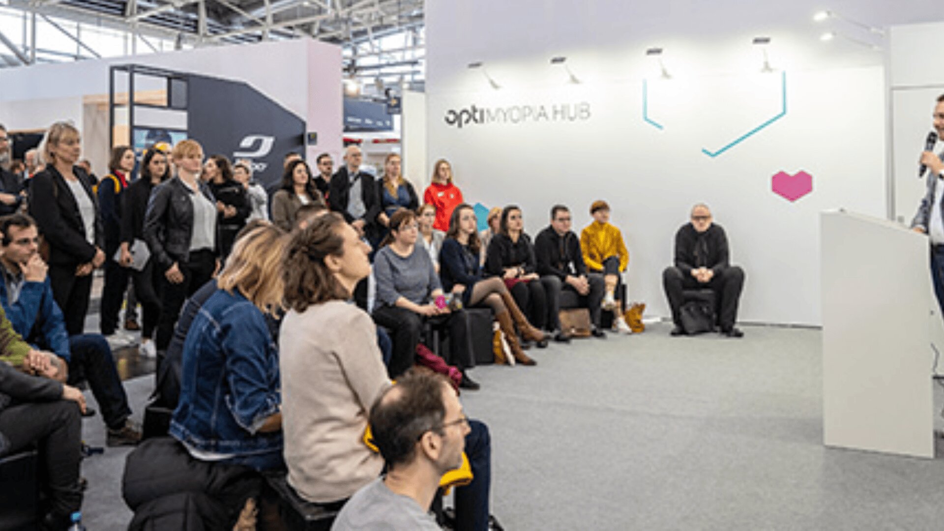 Eine Gruppe von Menschen sitzt und steht und hört aufmerksam einem Redner bei einer modernen Indoor-Veranstaltung oder Konferenz zu. Dahinter befindet sich eine weiße Wand mit der Aufschrift MOPA HUB und geometrischen Mustern.