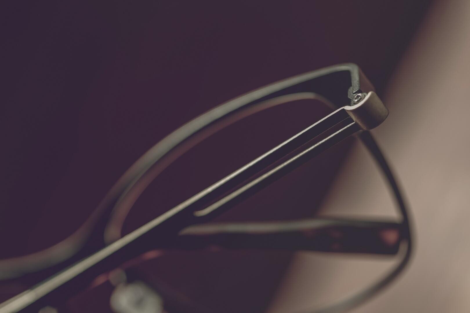 Nahaufnahme der Ecke einer Brille, wobei der Fokus auf den glatten, metallischen Bügeln und dem Scharnier vor einem leicht verschwommenen, dunklen Hintergrund liegt.
