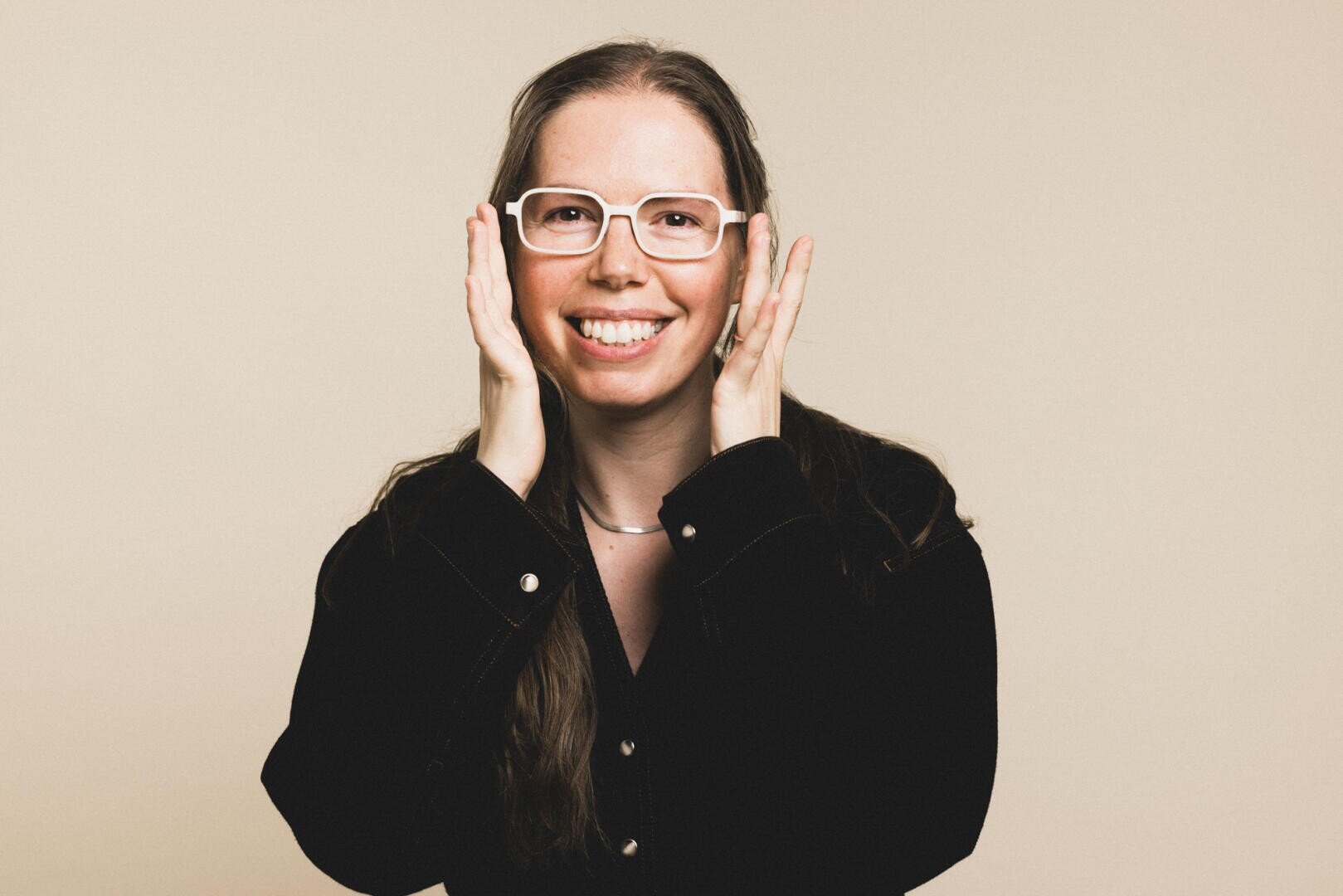 Eine lächelnde Person mit langen braunen Haaren, die eine weiße Brille und ein schwarzes Hemd trägt, berührt mit ihren Händen beide Seiten ihres Gesichts vor einem schlichten beigen Hintergrund.