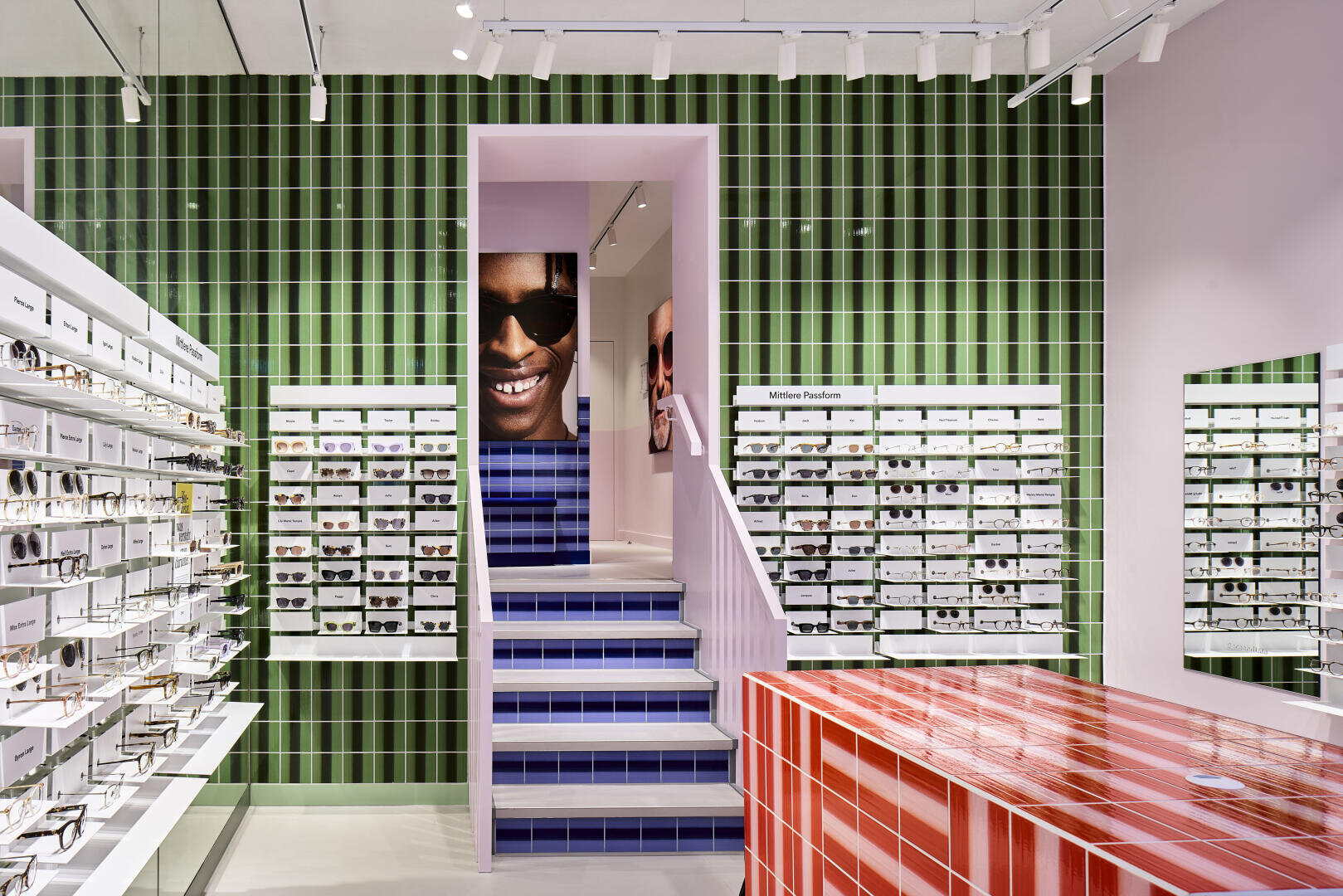 Ein modernes Brillengeschäft mit grünen vertikalen Kachelwänden, weißen Brillenregalen, einem rot gestreiften Tresen, einer blau gekachelten Treppe und einem großen Foto mit einem lächelnden Gesicht über der Treppe.