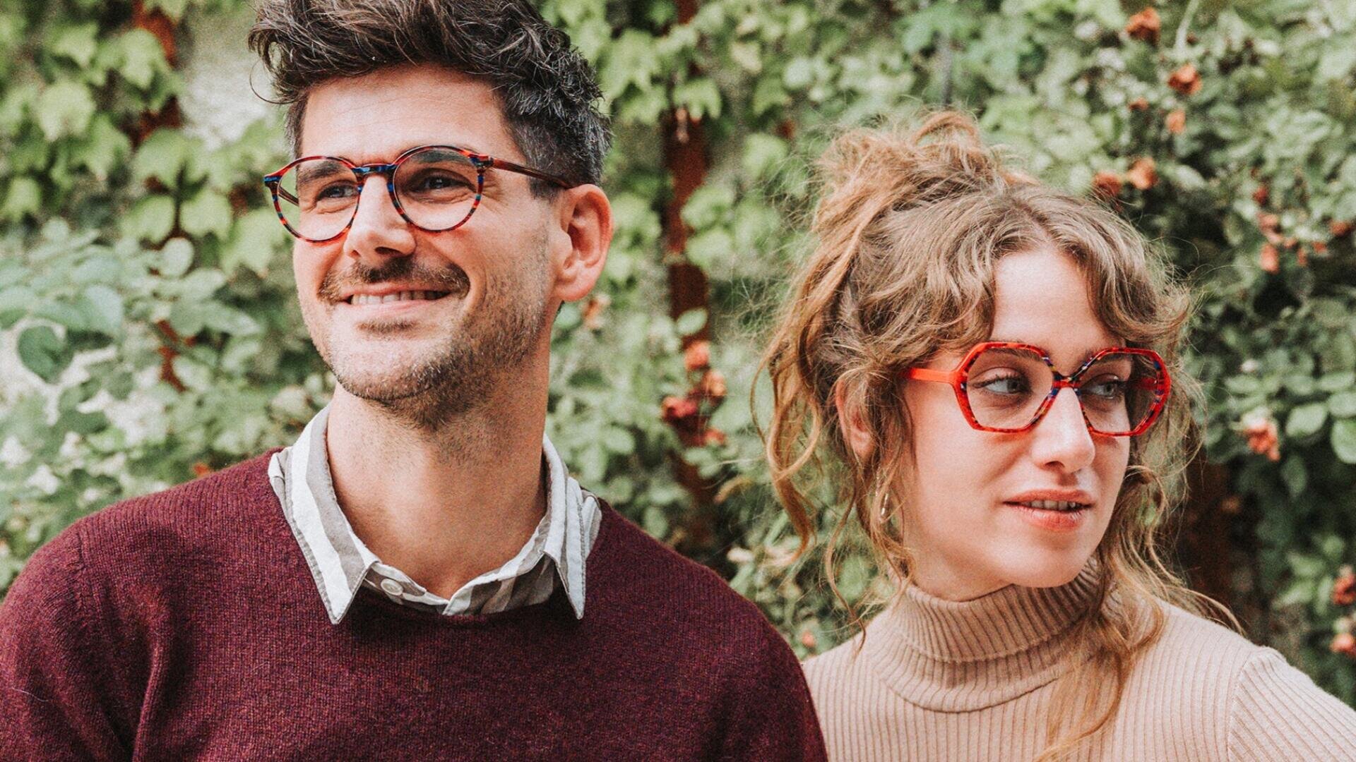 A man and a woman with glasses are standing outside in front of green plants. The man has short dark hair and is wearing a maroon sweater, while the woman has light brown hair in a messy bun and is wearing a beige turtleneck sweater.