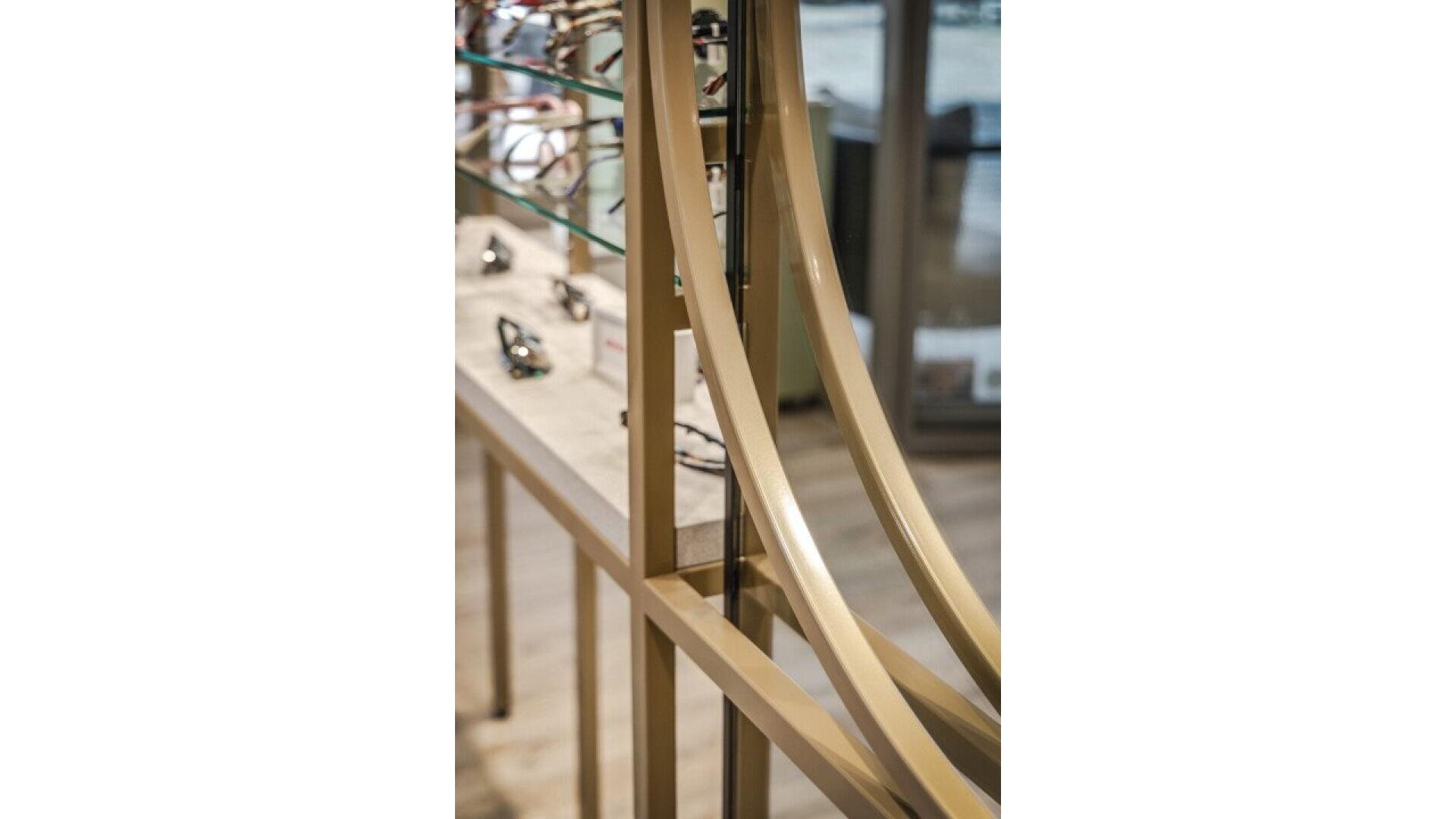 Close-up of a modern metal display stand with curved gold bars, glass shelves, and several pairs of eyeglasses arranged on the shelves in a well-lit store interior.