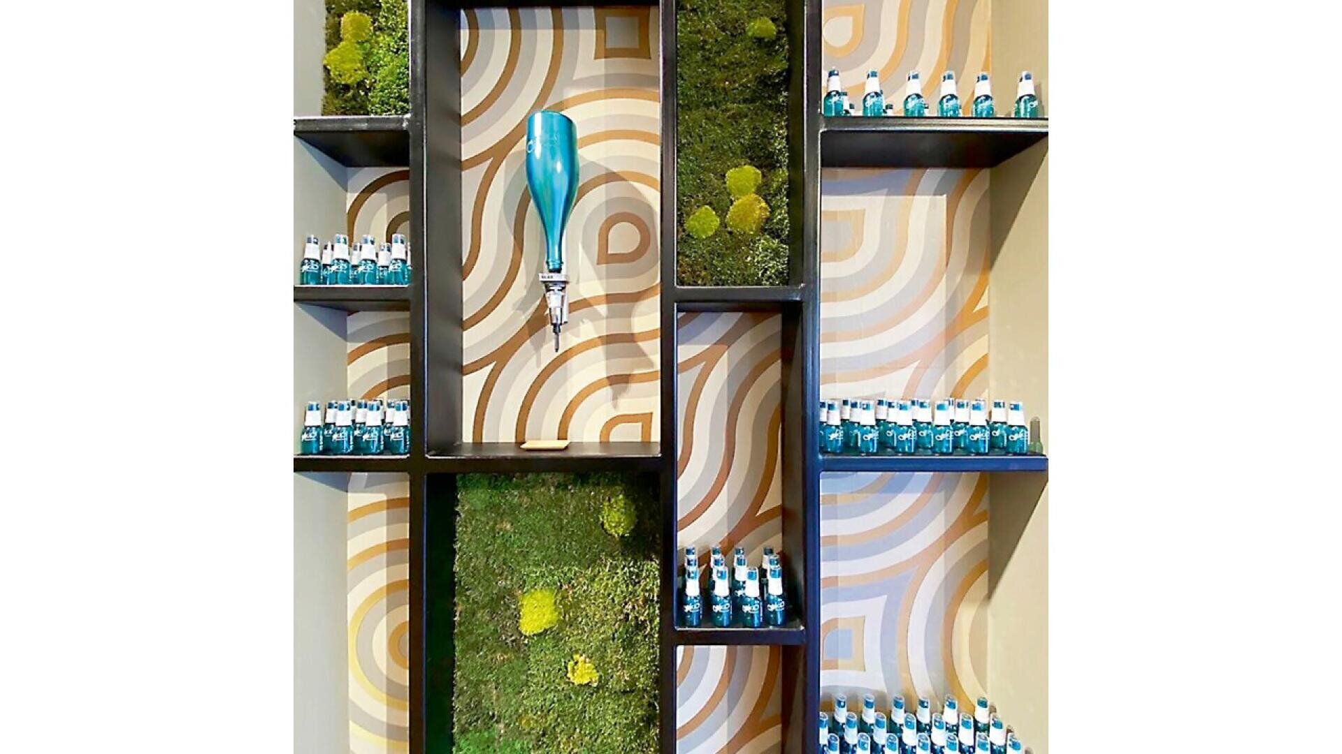 A wall display with geometric shelves, small teal bottles, patches of green moss, and a blue beverage tap against a retro-patterned background of curved tan and orange lines.