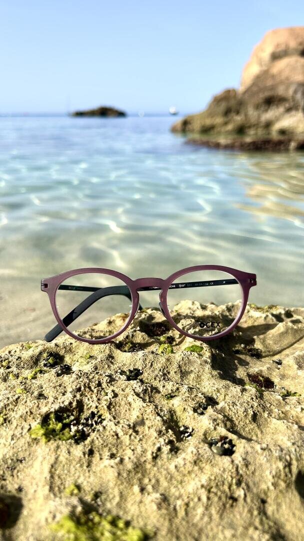 Eine lilafarbene Brille ruht auf einem moosbewachsenen Felsen am Rande eines klaren, flachen Wassers mit weiteren Felsen im Hintergrund unter einem sonnigen, blauen Himmel.