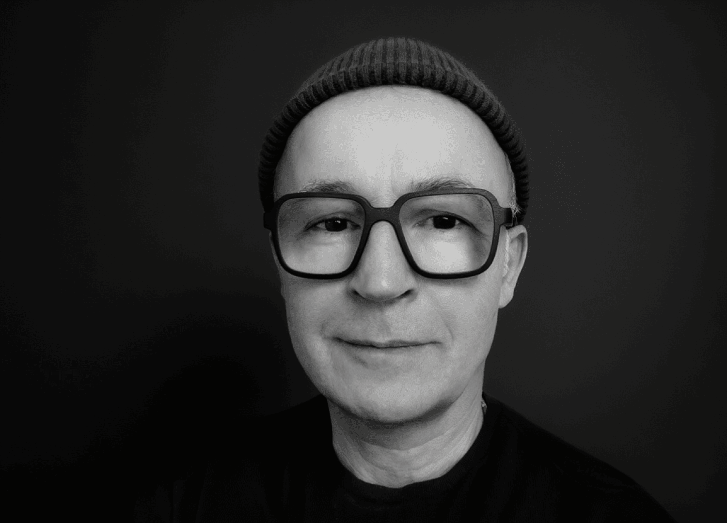 Eine Person, die eine große, dunkel gerahmte Brille und eine Strickmütze trägt, lächelt leicht vor einem schlichten, dunklen Hintergrund in diesem Schwarz-Weiß-Porträt.