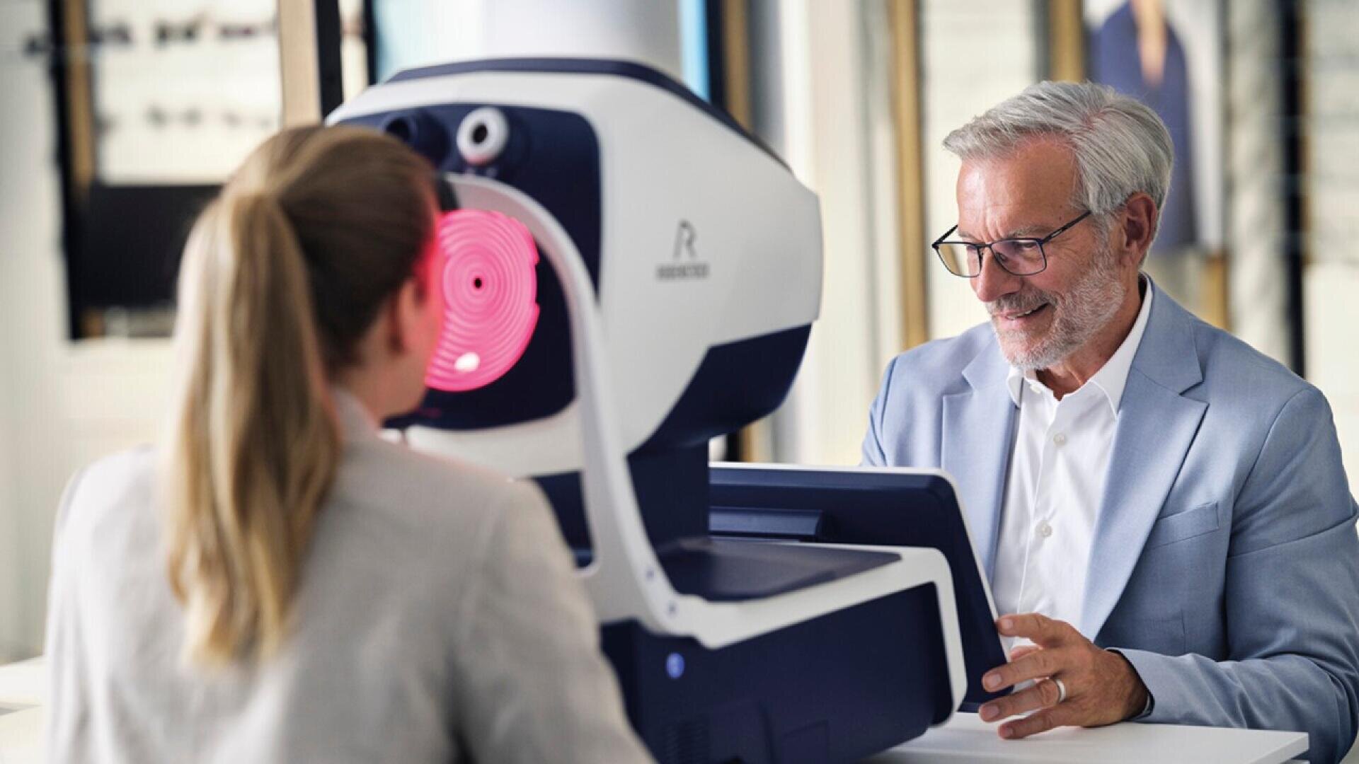An older man in a light suit smiles while operating an eye exam machine for a woman with a ponytail, who is seated with her face in the device emitting red concentric circles.