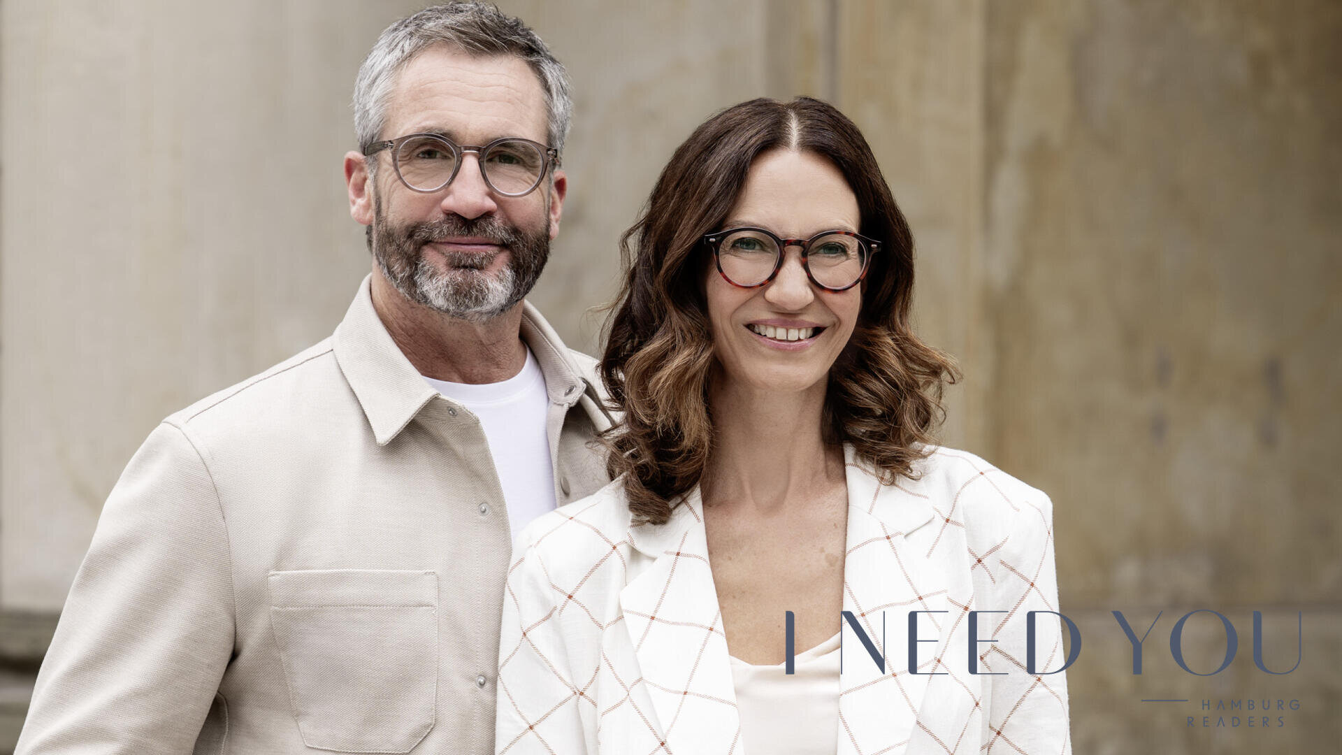 Ein lächelnder Mann mit grauem Haar und Bart steht neben einer lächelnden Frau mit gewelltem braunem Haar. Beide tragen eine Brille und helle Jacken. Der Text I NEED YOU HAMBURG READERS ist in der unteren rechten Ecke zu sehen.
