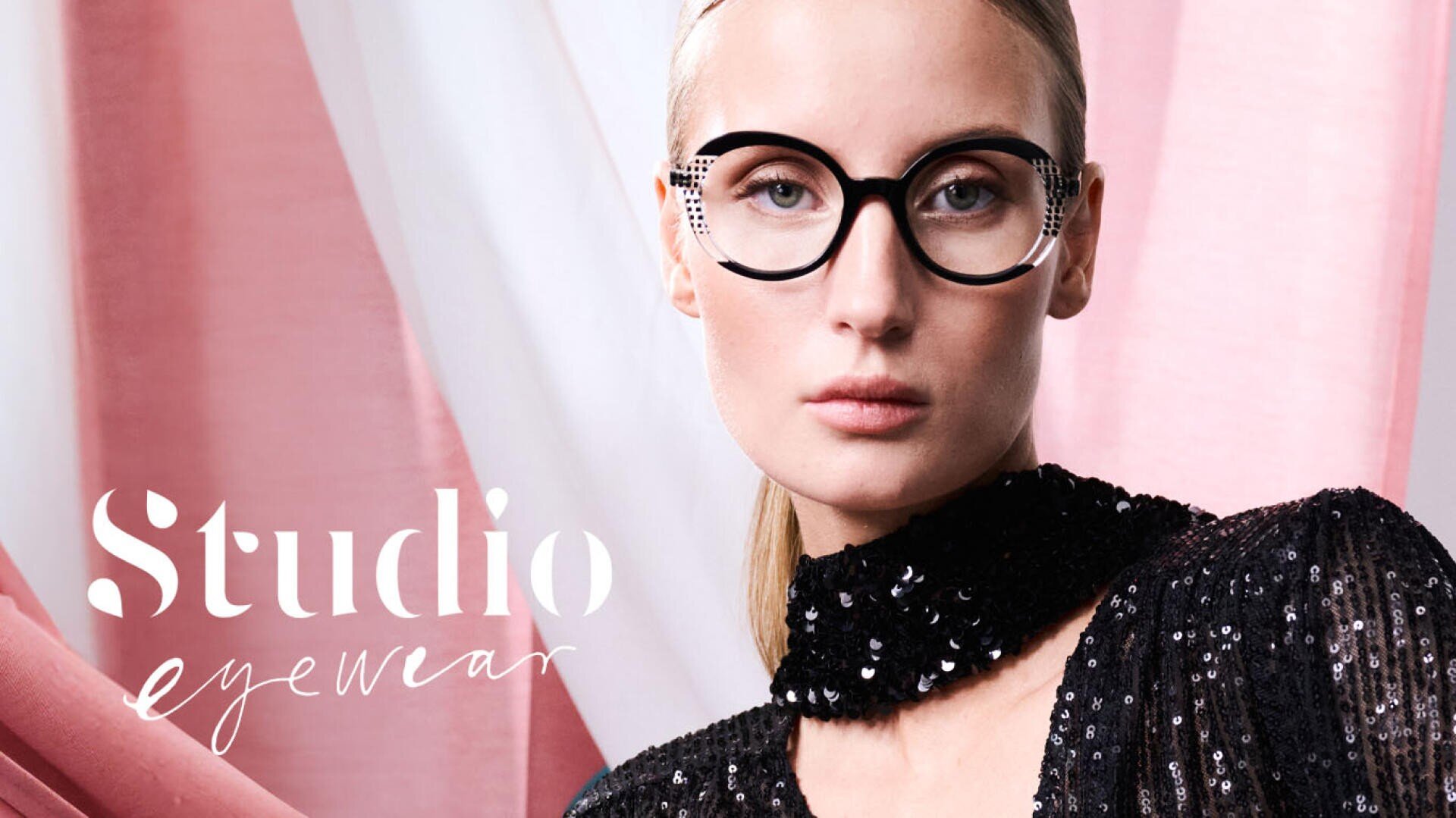 Eine Frau mit großer, runder schwarzer Brille und schwarzem Pailletten-Outfit posiert vor einem rosa und weiß drapierten Stoff. Der Text "Studio eyewear" erscheint auf der linken Seite des Bildes.