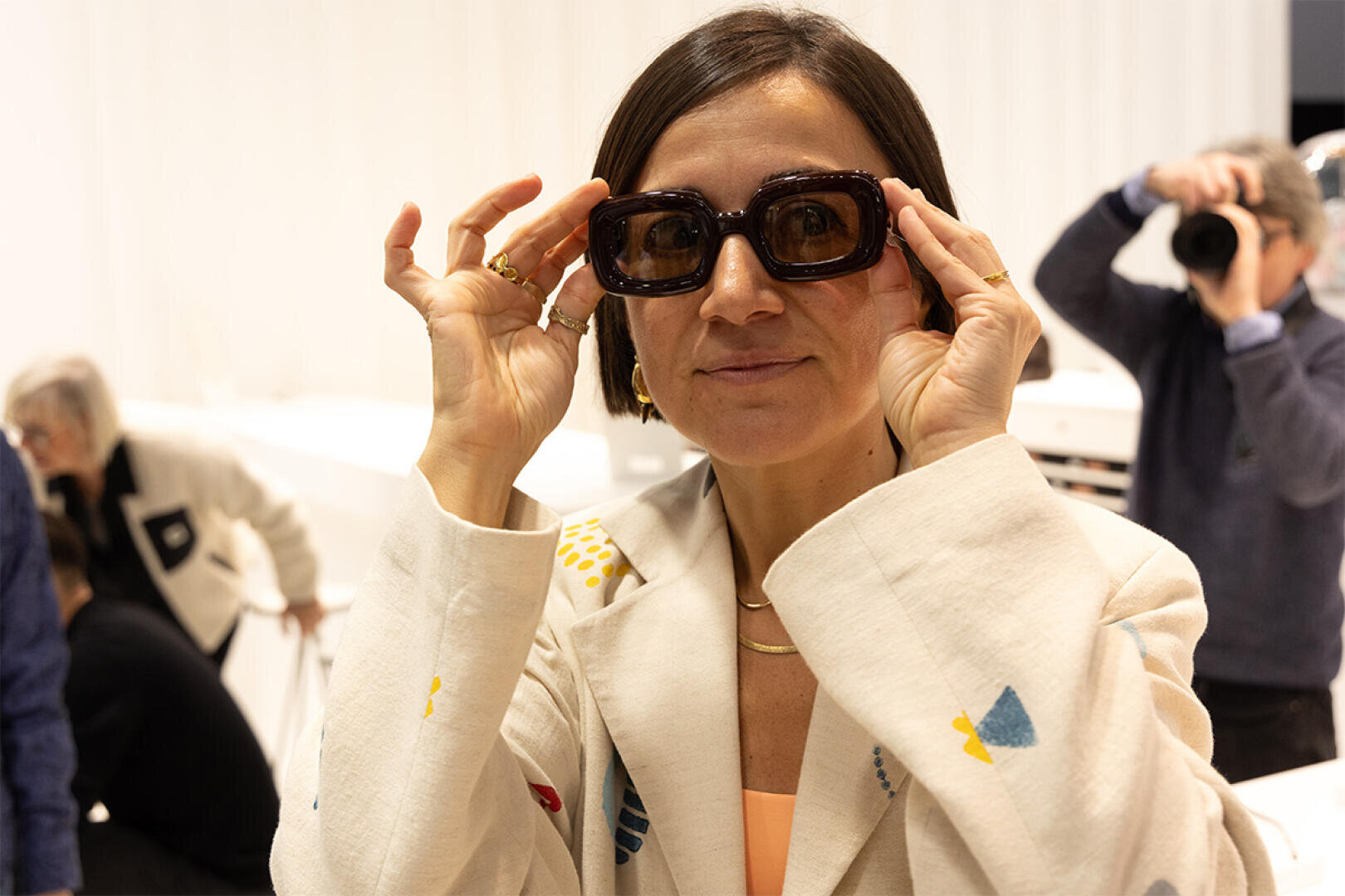Eine Frau in einem hellen Blazer mit bunten Mustern rückt eine große, dunkle Sonnenbrille zurecht und lächelt. Im Hintergrund macht eine Person ein Foto, und weitere Personen sind in einem gut beleuchteten Innenraum anwesend.
