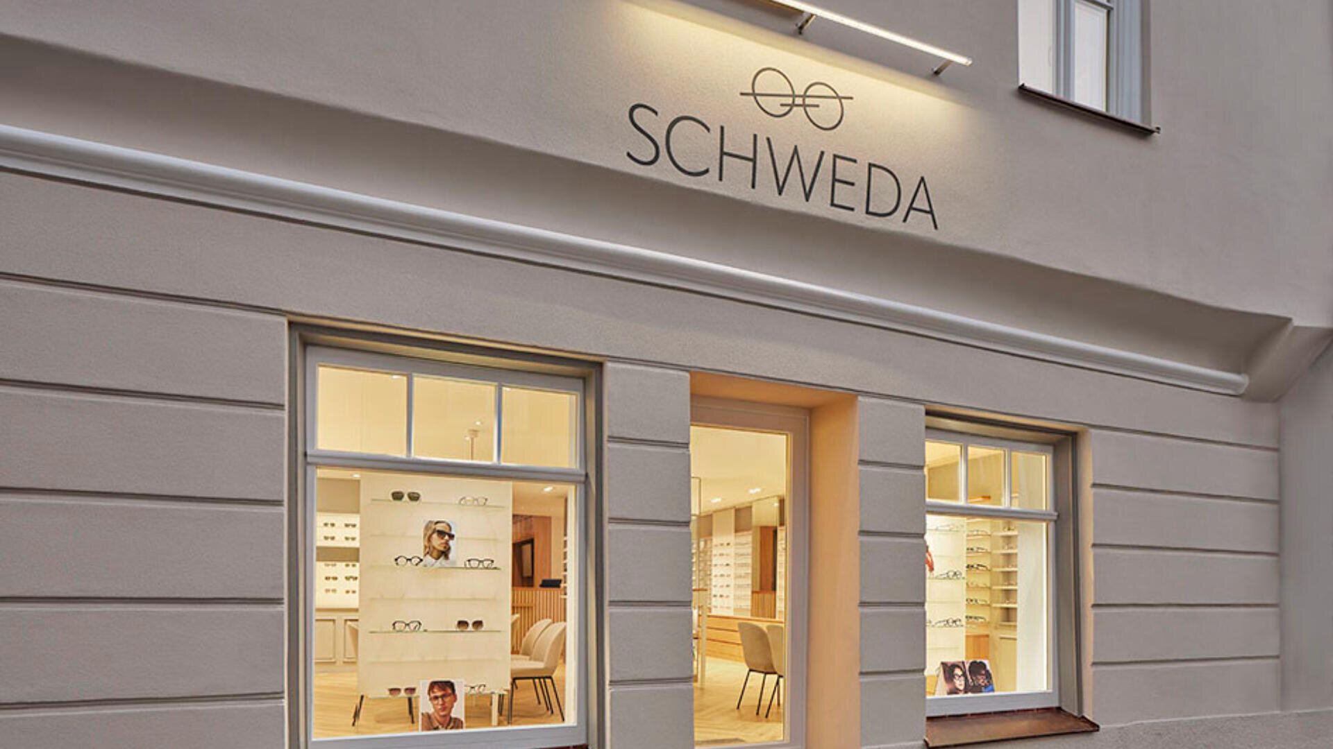 Straßenansicht des Brillengeschäfts SCHWEDA mit großen Schaufenstern und einem hellen, modernen Innenraum, der durch die Glasfronten sichtbar ist. Über dem Eingang sind das SCHWEDA-Logo und die Beschilderung angebracht.