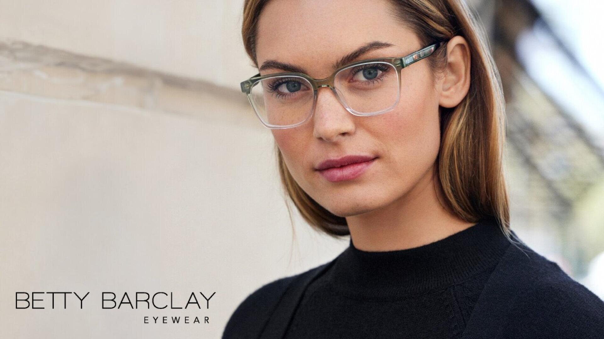 A woman with light brown hair and clear-framed glasses looks confidently at the camera, wearing a black top. The image features the text Betty Barclay Eyewear in the bottom left corner.