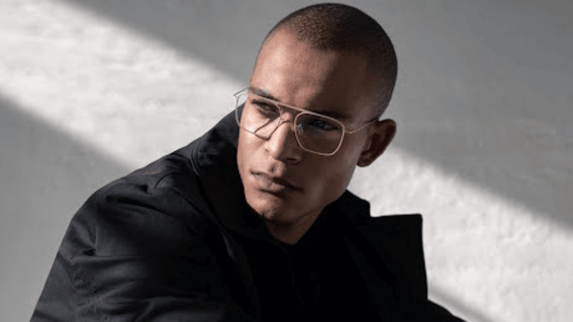 Ein Mann mit kurzen Haaren und klarer Brille, der eine dunkle Jacke trägt, blickt bei sanftem, natürlichem Licht zur Seite, mit Schatten auf einer weißen Wand hinter ihm.