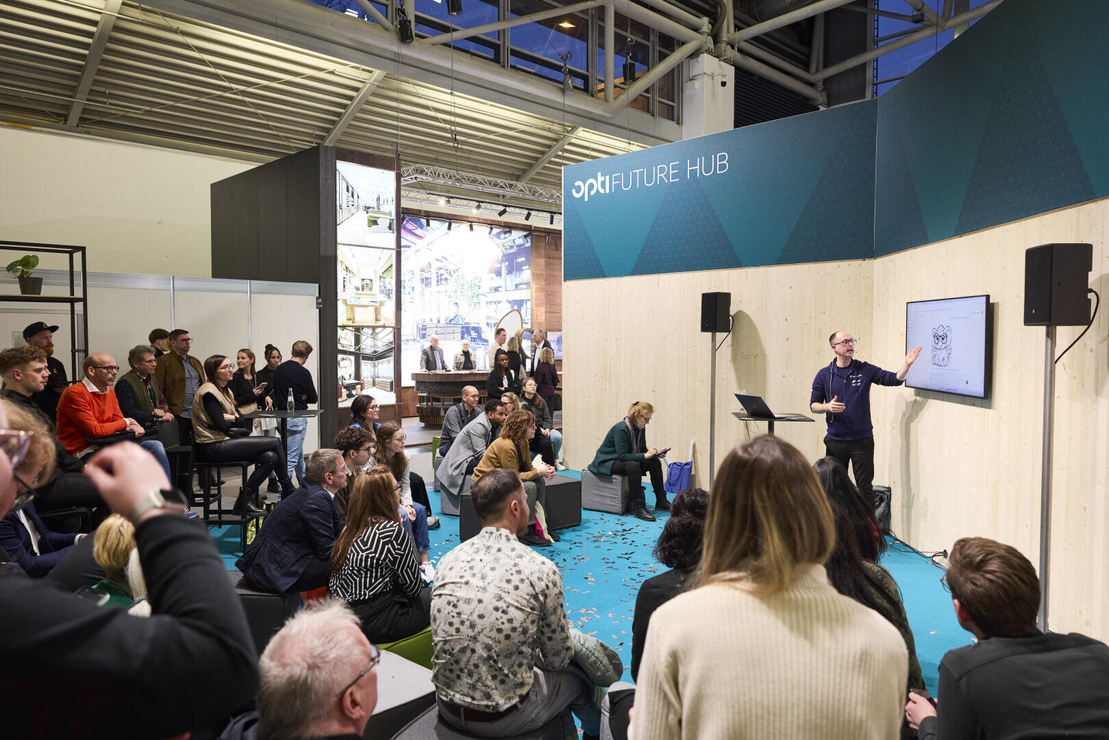 Ein Redner spricht zu einem aufmerksamen Publikum in einem modernen Veranstaltungsraum mit der Aufschrift FUTURE HUB, auf dessen Bildschirm eine Cartoon-Grafik zu sehen ist. Die Menschen sitzen und stehen, hören zu und machen sich Notizen.