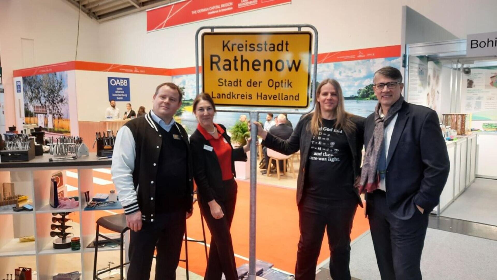 Four people stand indoors around a large yellow sign reading Kreisstadt Rathenow, Stadt der Optik, Landkreis Havelland. Booths and displays are visible in the background, suggesting a trade fair or exhibition setting.