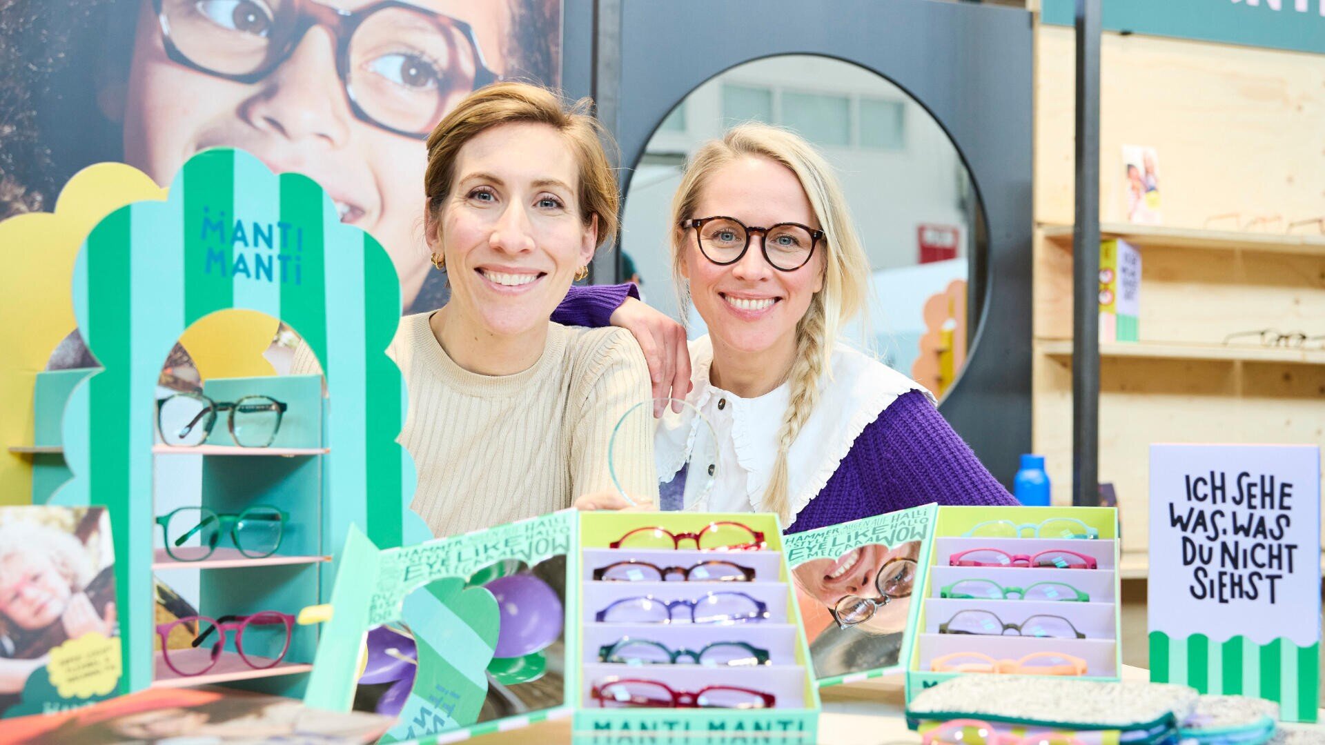 Zwei lächelnde Frauen mit Brille stehen an einem Innenstand hinter einem farbenfrohen Display aus Brillen und Werbeschildern. Auf einem Schild auf Deutsch steht: „Ich sehe was, was du nicht siehst.“.