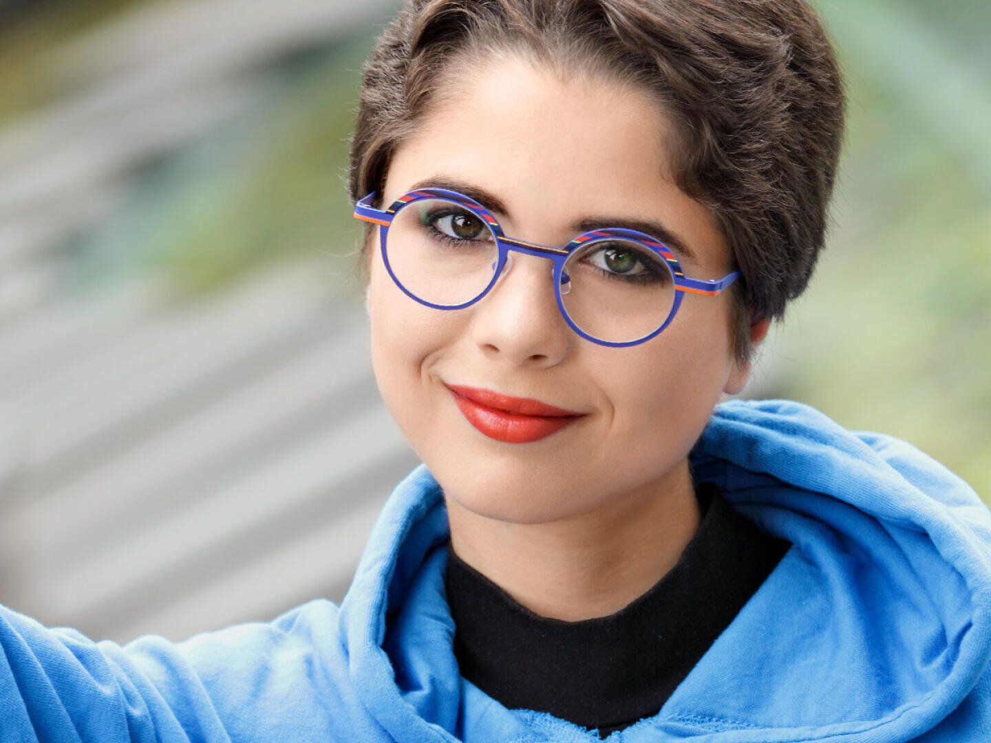 Eine Person mit kurzen braunen Haaren, die eine runde blaue Brille, einen hellblauen Kapuzenpullover und roten Lippenstift trägt, lächelt in die Kamera. Der Hintergrund ist mit Grün- und Grautönen verschwommen.