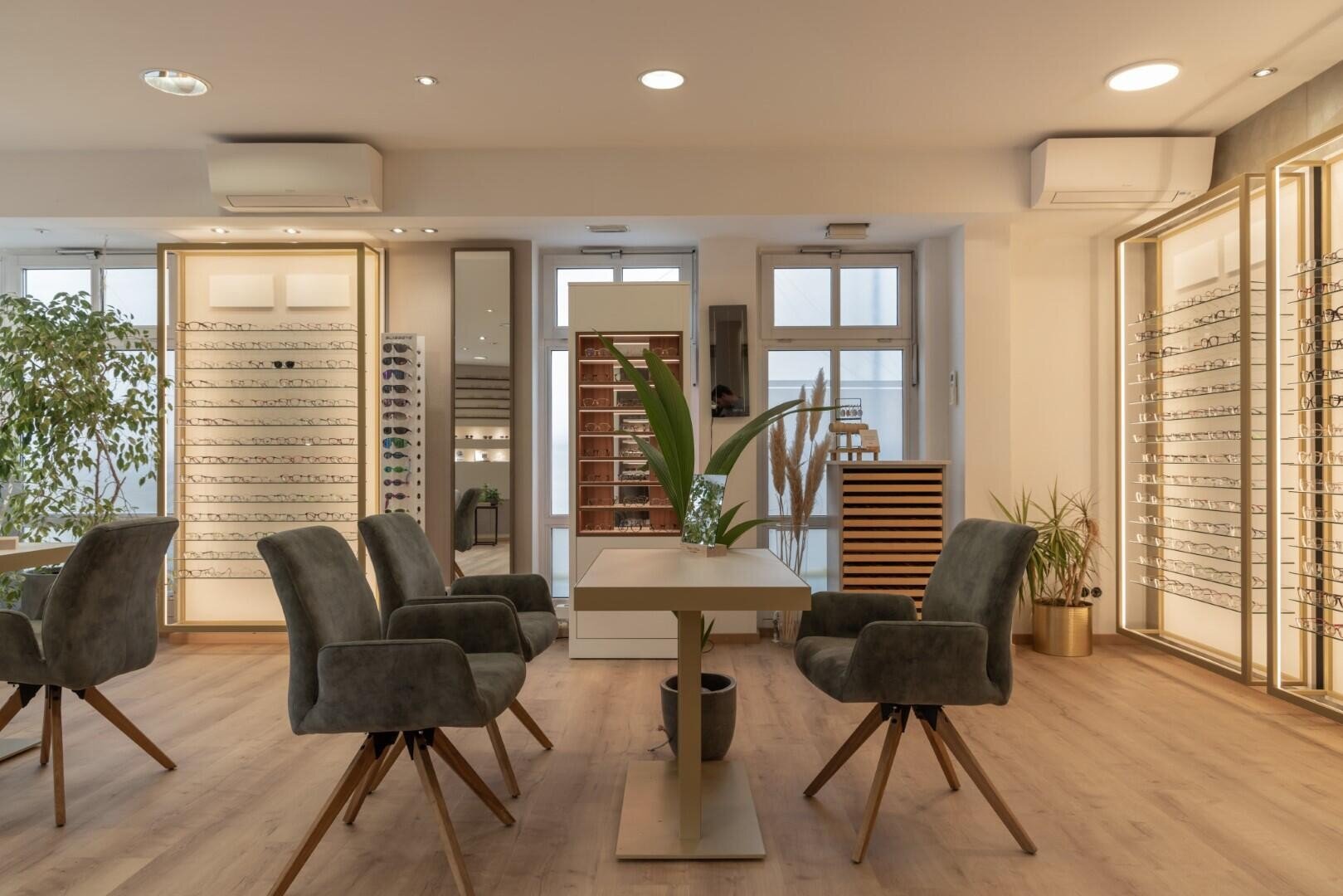 Ein modernes Optikergeschäft mit mehreren grauen Samtstühlen um Tische, Regalen voller Brillen, großen Fenstern, Pflanzen und Holzboden. Der Raum ist gut beleuchtet und übersichtlich.