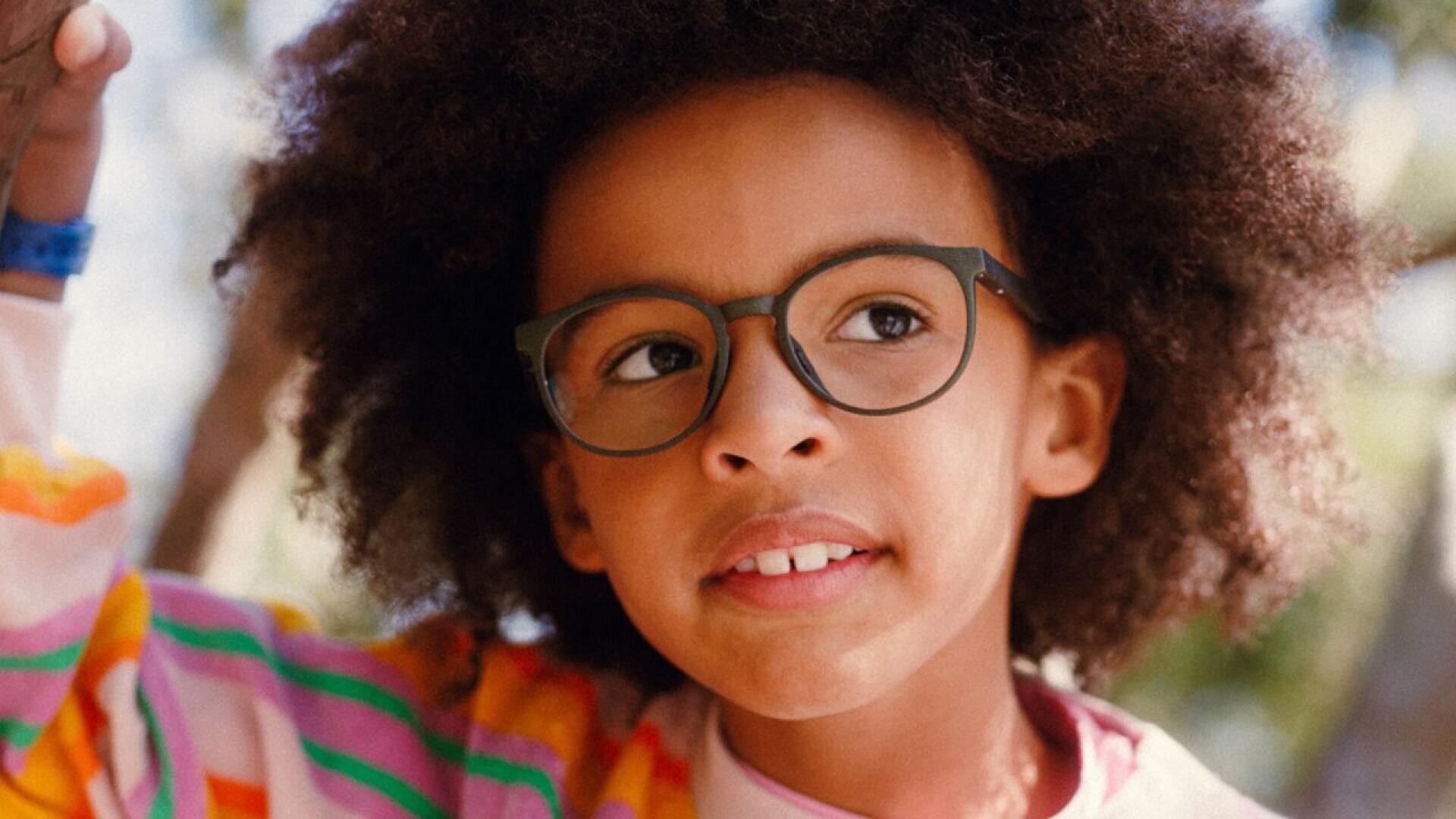 A child with curly hair and glasses is wearing a colorful striped sweater and smiling slightly while looking to the side outdoors.