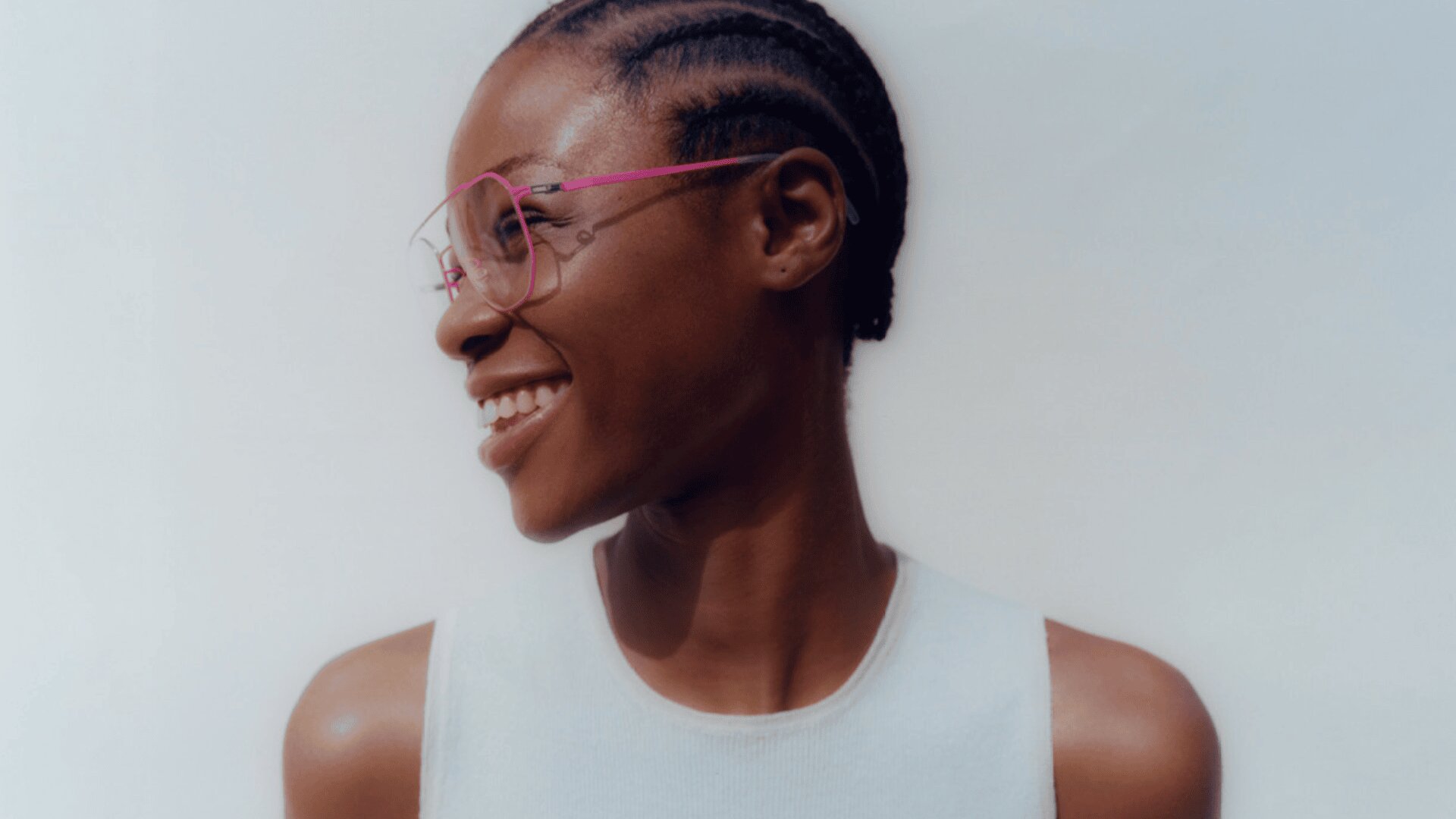 A person with short, braided hair smiles while looking to the left. They wear large, stylish, clear-framed glasses with pink arms and a white sleeveless top. The background is plain and light-colored.