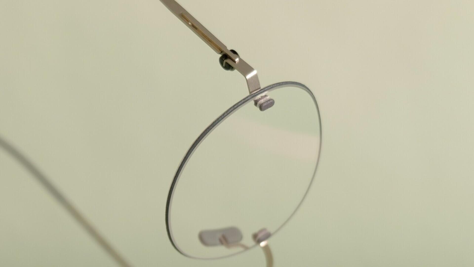 Close-up of a single round eyeglass lens with a thin metal frame and nose pad, set against a plain, light background.