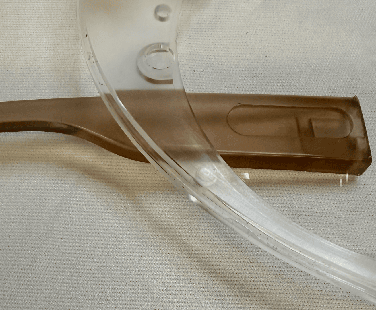 Nahaufnahme eines durchsichtigen Kunststoffvisiers, das einen braunen Kunststoffgriff überlappt; beide liegen auf einer hellen, strukturierten Stoffoberfläche.
