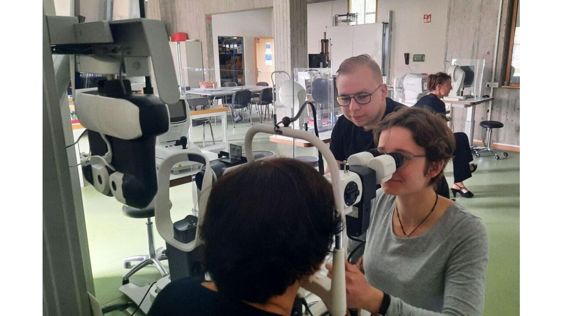 Three people in a clinical or educational setting use ophthalmology equipment; one person sits at the machine while another observes, and a third person looks on in the background.
