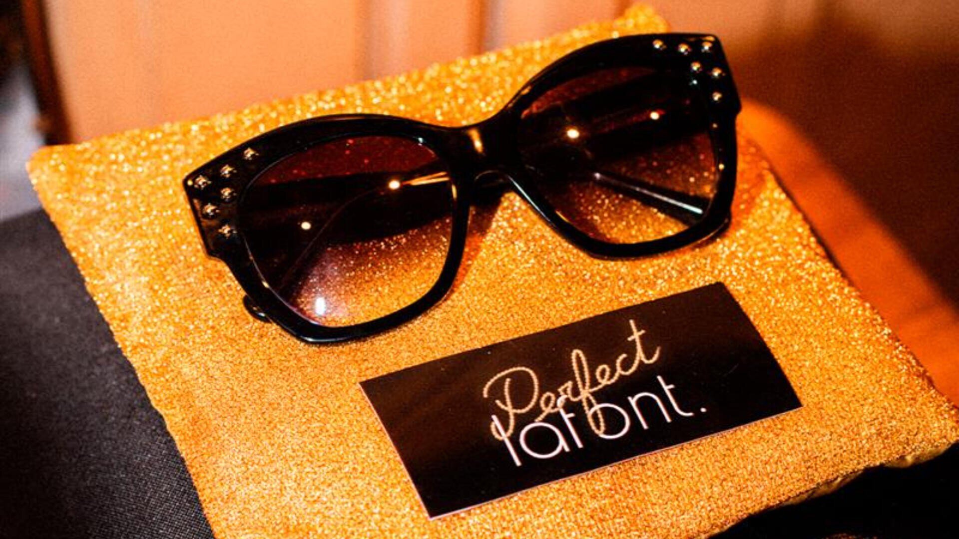 Große, schwarze Sonnenbrillen mit Verlaufsgläsern liegen auf einem glitzernden Goldstoff neben einer schwarzen Karte mit der Aufschrift „Perfect lafânt“.