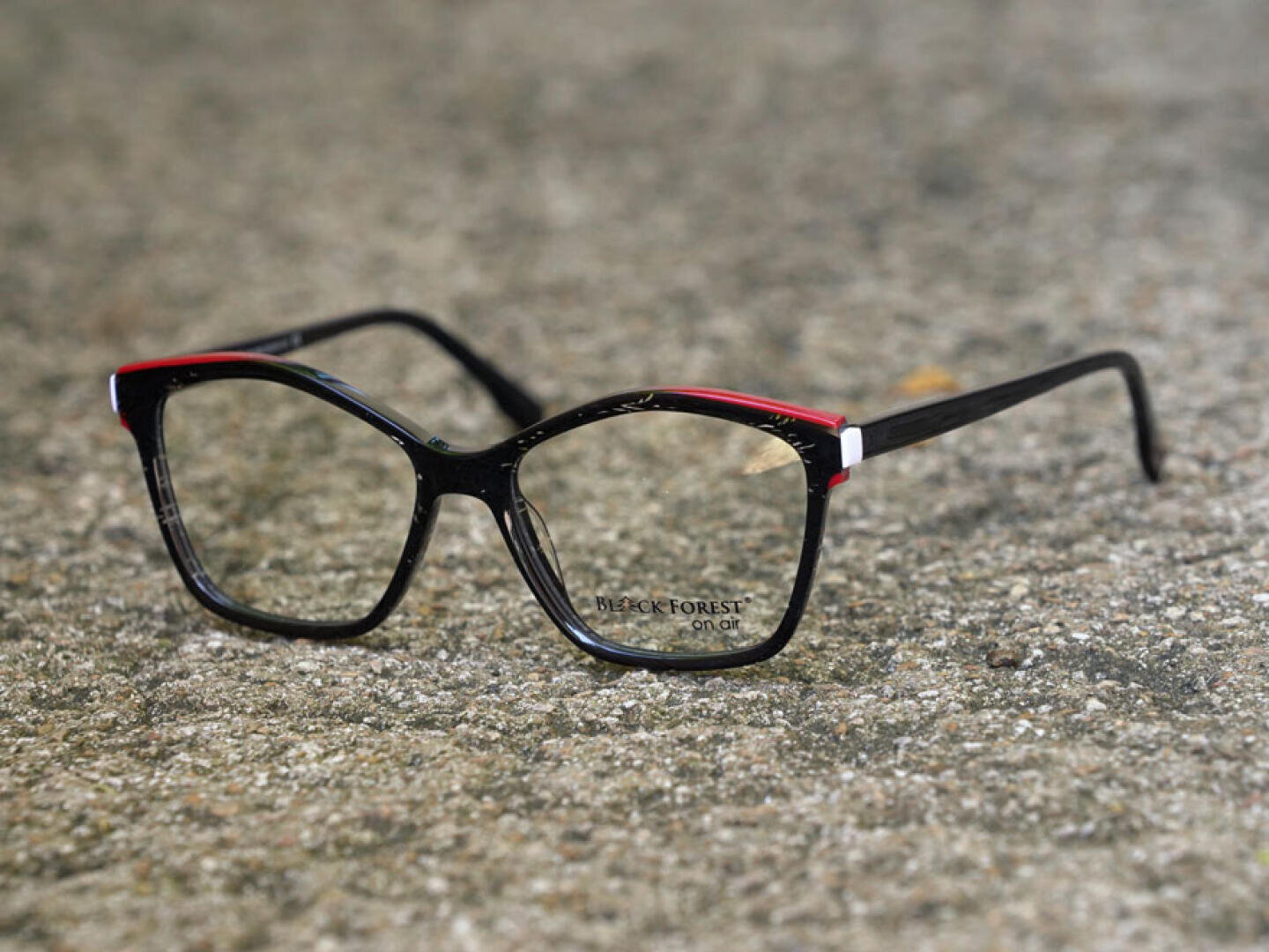Eine schwarz umrandete Brille mit dezenten roten Akzenten auf der Oberseite, die auf einer strukturierten grauen Oberfläche ruht. Auf dem linken Glas steht der Markenname BLACK FOREST.
