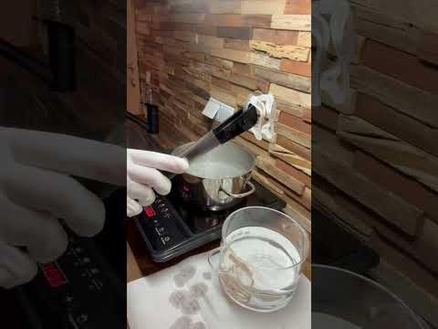 Eine Person, die einen weißen Handschuh trägt, taucht mit einer Zange einen weißen Gegenstand in einen Topf mit kochendem Wasser auf einem Herd. Eine Schale mit weißem Pulver und einige Papiertücher stehen in der Nähe auf dem Tresen.