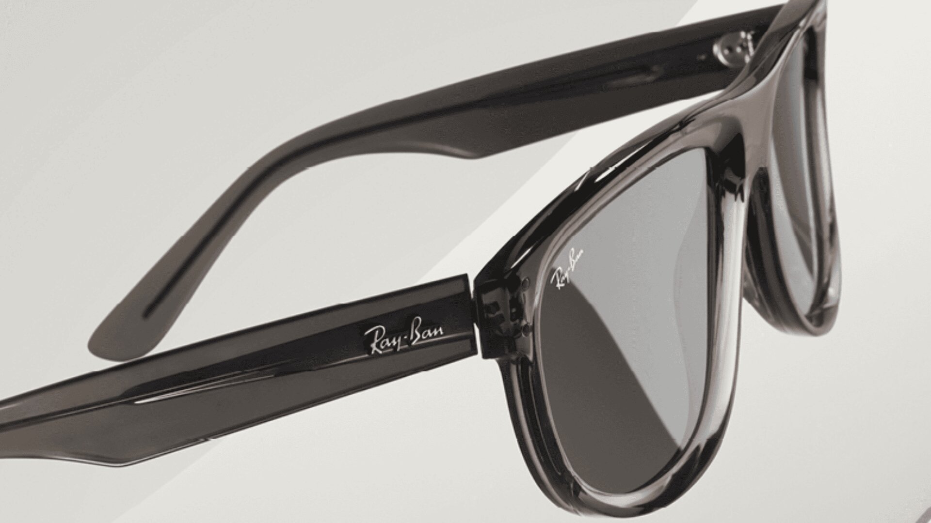 Nahaufnahme einer schwarzen Ray-Ban-Sonnenbrille mit dunklen Gläsern, die das Ray-Ban-Logo auf dem Bügel und den Gläsern zeigt, vor einem hellen, neutralen Hintergrund.