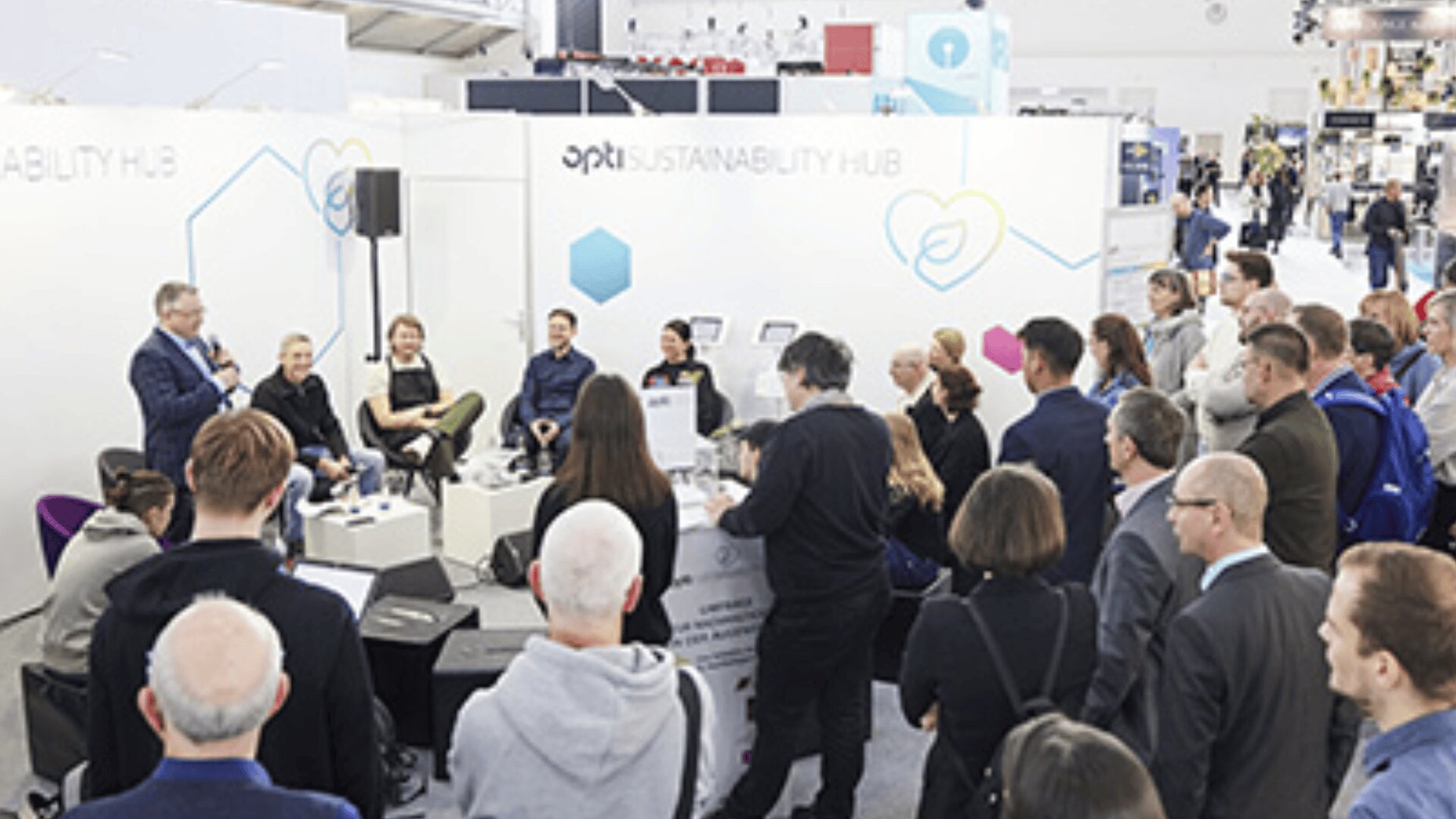 Eine Gruppe von Menschen hört einer Podiumsdiskussion auf einer Konferenz aufmerksam zu. Die Diskussionsteilnehmer sitzen auf Stühlen vor einem weißen Stand mit der Aufschrift "Sustainability Hub", während die Teilnehmer in der Nähe stehen oder sitzen.