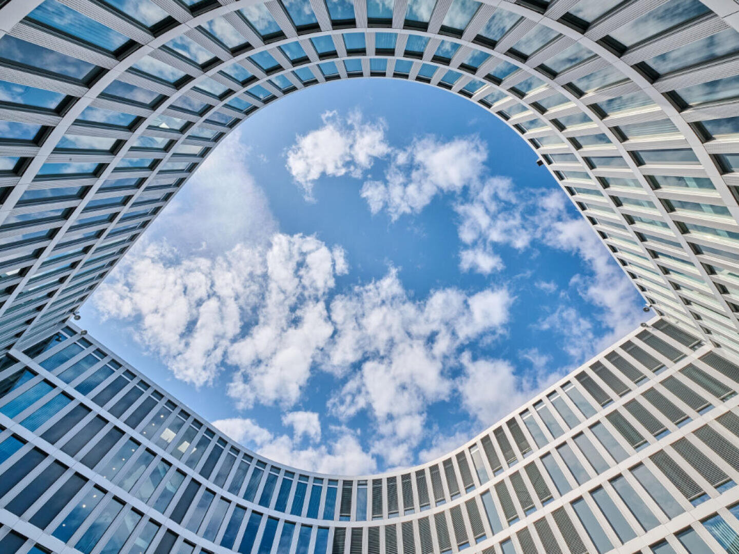 A modern building with curved glass windows surrounds an open oval space, above which a blue sky with scattered clouds can be seen. The photo was taken from the ground, looking straight up.