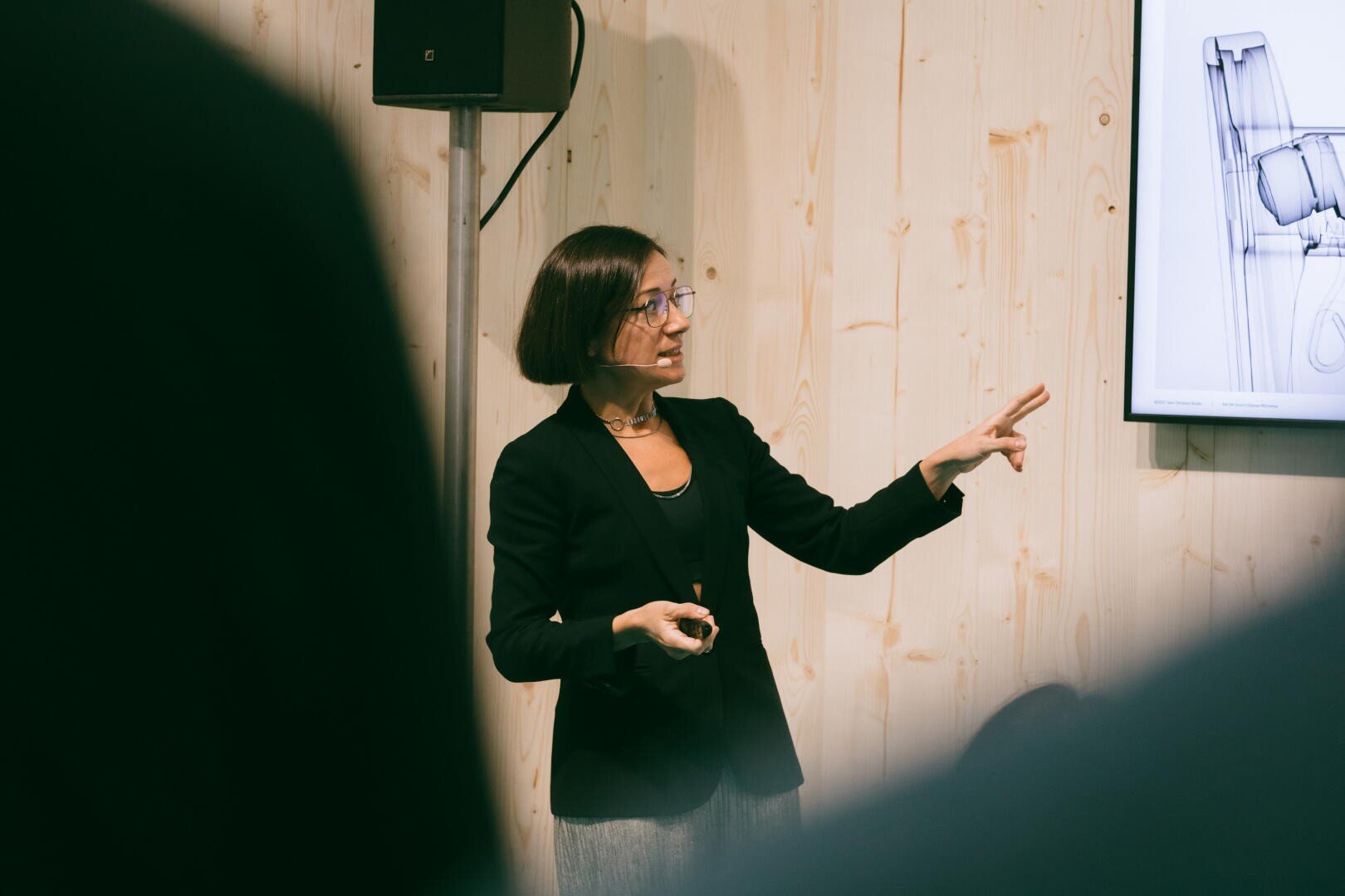 Eine Frau in schwarzem Blazer und mit Brille hält einen Vortrag und zeigt auf einen Bildschirm, auf dem eine technische Zeichnung zu sehen ist, mit einer hellen holzgetäfelten Wand im Hintergrund.