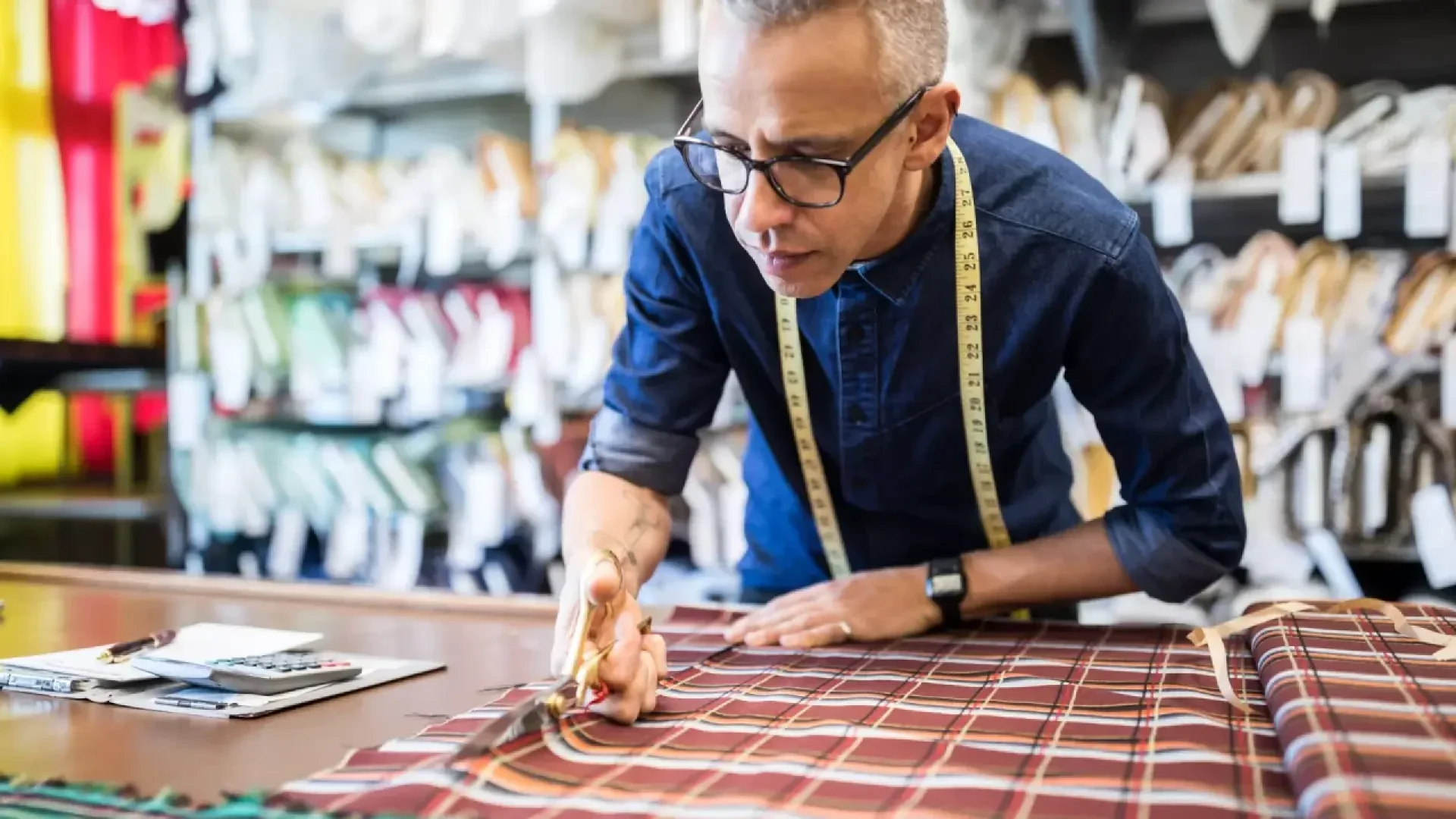 A person wearing glasses and a tape measure around their neck cuts plaid fabric with large scissors on a work table in a tailor shop. Rolls of fabric and sewing tools are visible in the background.