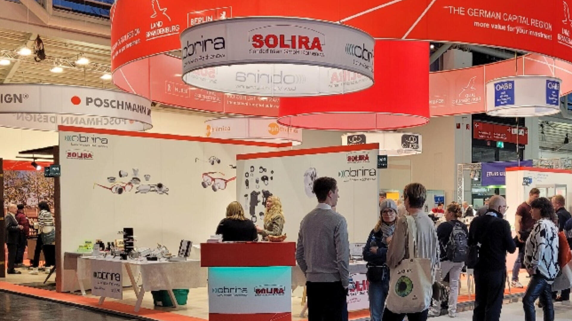 Ein belebter Messestand mit interagierenden Menschen, Produktpräsentationen, Broschüren auf Tischen und großen rot-weißen Schildern über den Köpfen, auf denen der Name SOLIRA und andere Markenzeichen zu sehen sind.