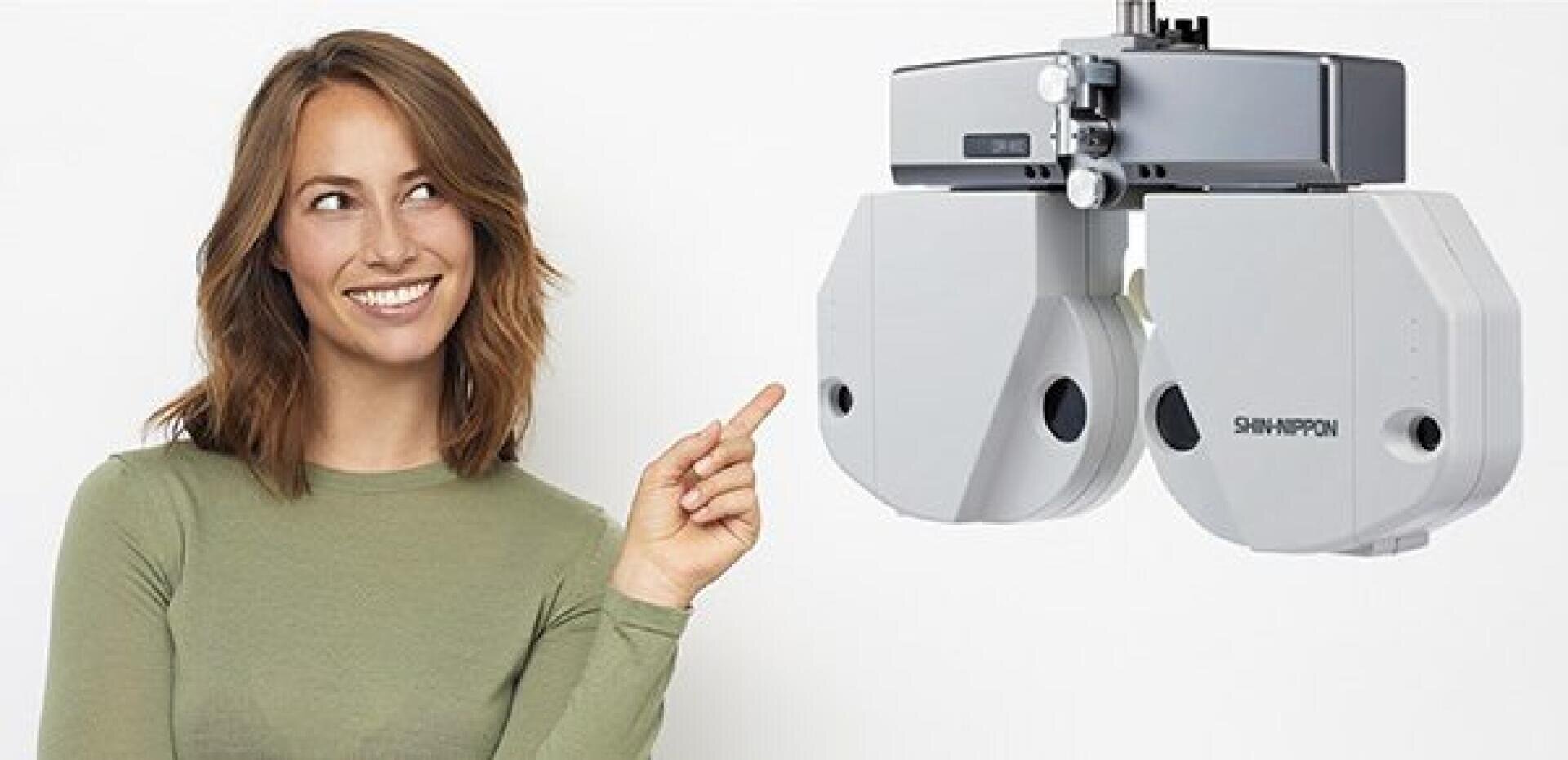 Eine Frau mit schulterlangem braunem Haar und einem grünen Oberteil lächelt und zeigt auf ein modernes augenärztliches Gerät mit der Aufschrift „SHINNIPPON“ vor einem schlichten weißen Hintergrund.